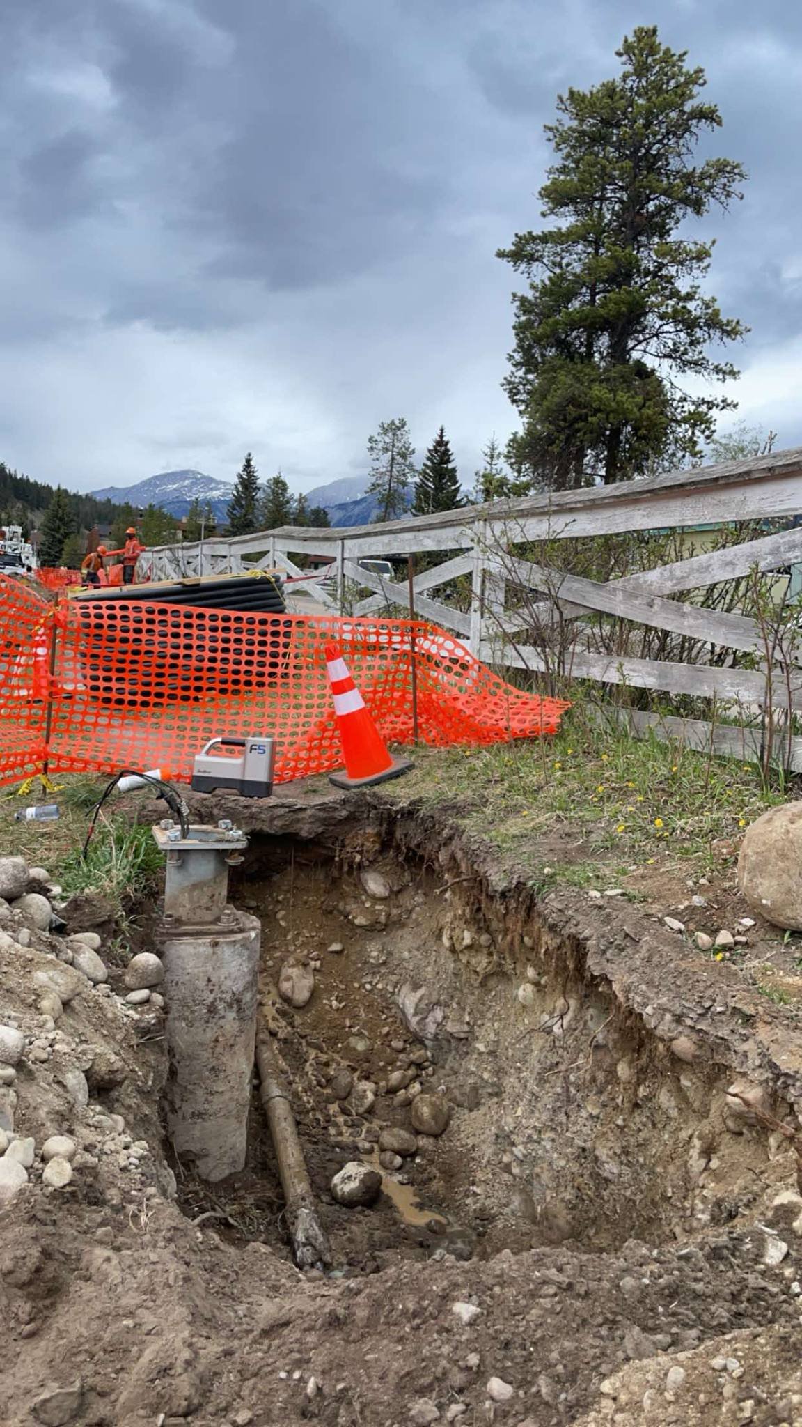 An outdoor construction site with a large hole in the ground, orange safety fencing, and traffic cones. There are workers in the distance, along with construction tools and equipment. The background features trees and mountains under cloudy skies.