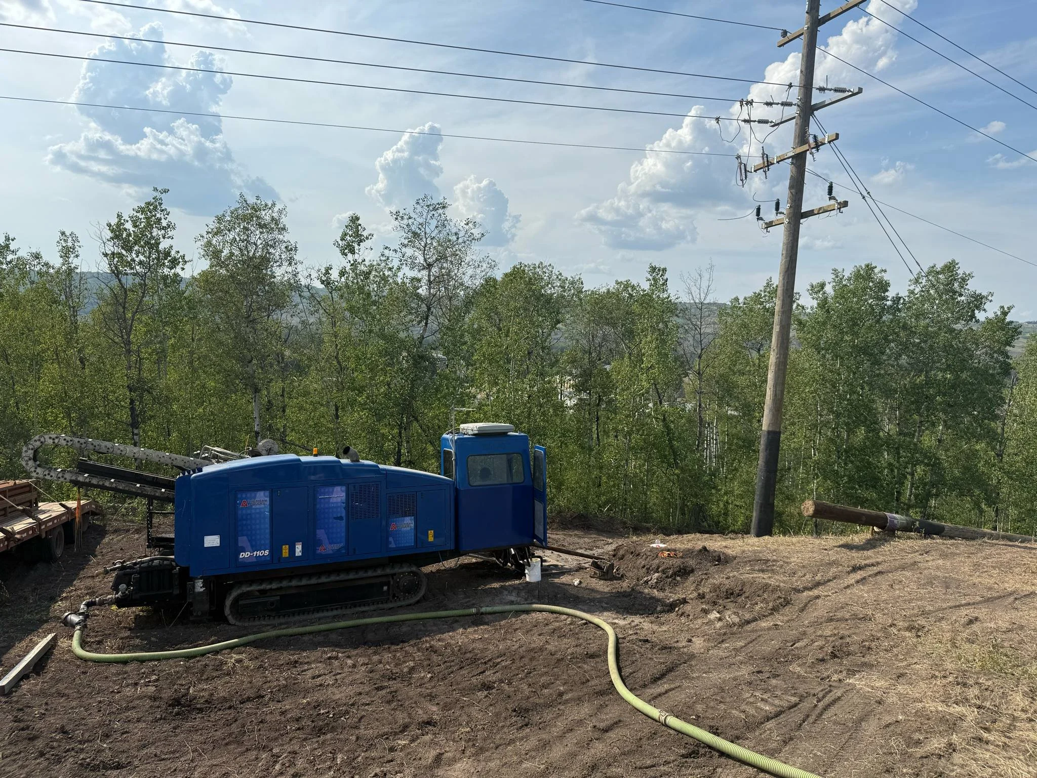 A large blue industrial machine on tracks next to a fallen utility pole and power lines in a rural area with trees and a partly cloudy sky.