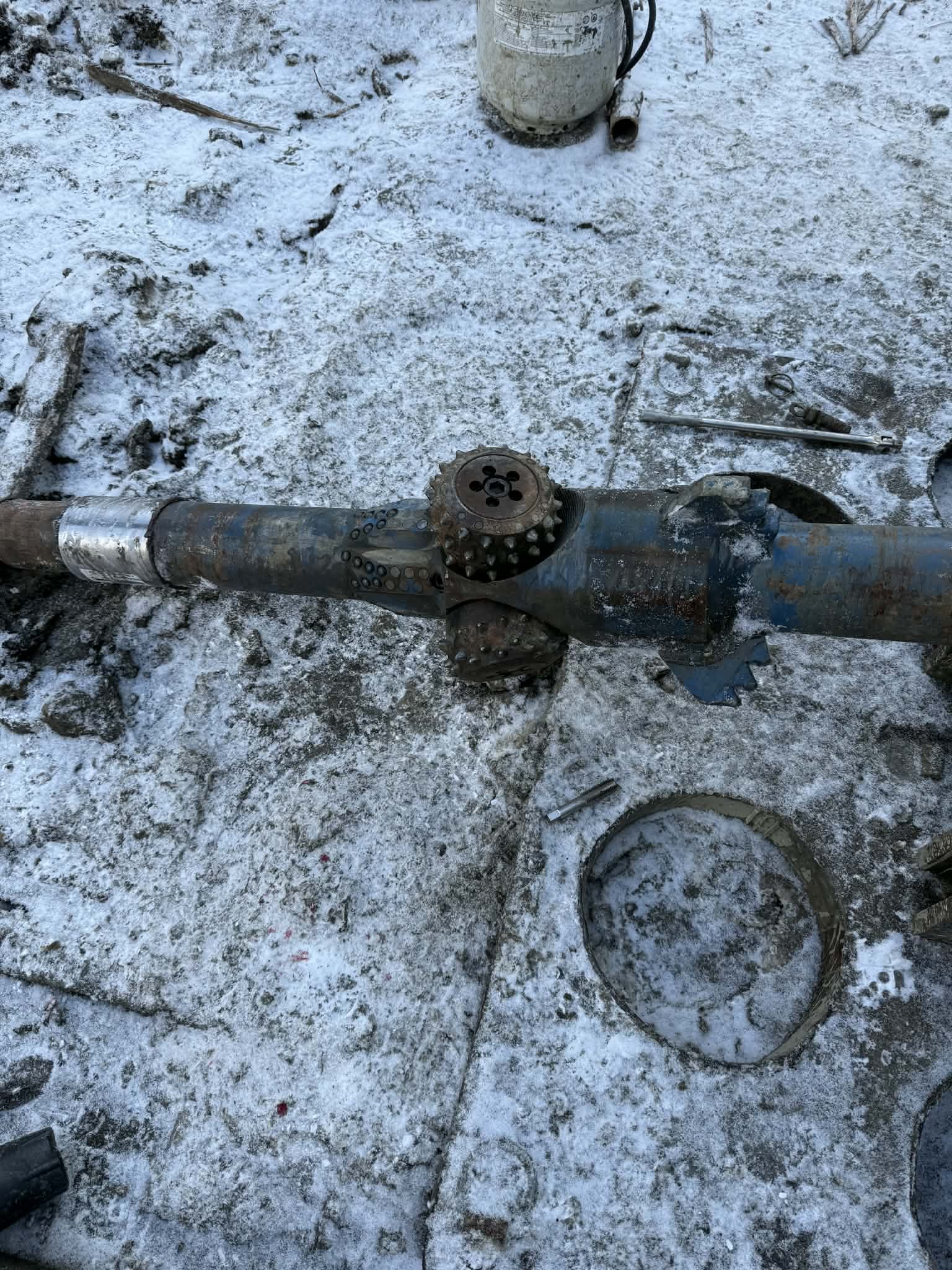 Disassembled mechanical part on snowy ground with various tools and a container in the background.