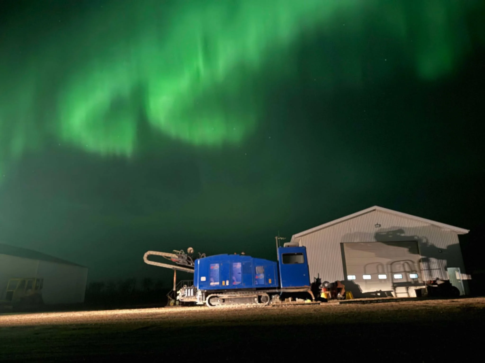 Northern lights in the sky above a blue horizontal directional drill and a white industrial building at night. American Augers DD110