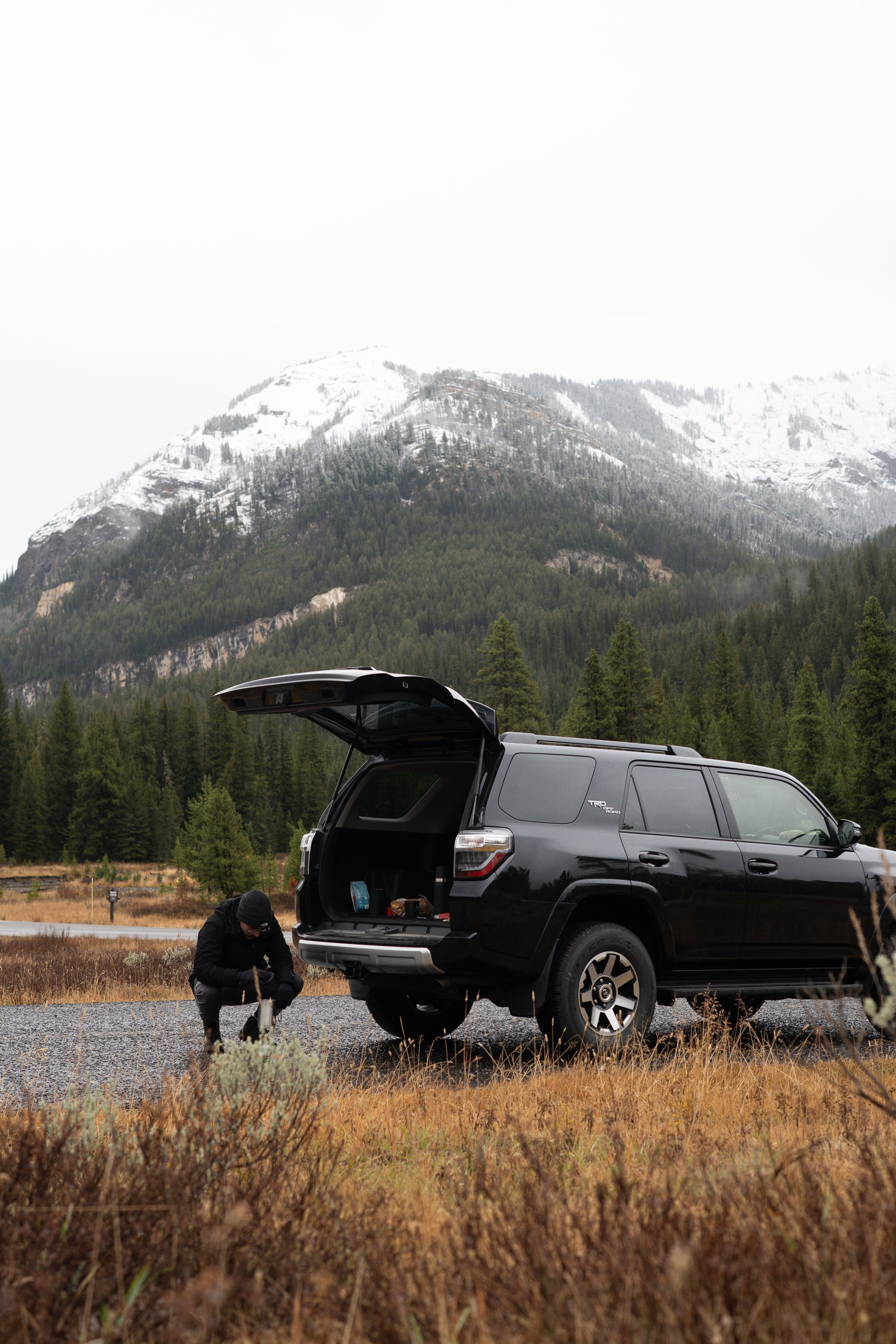 A person crouching near the open trunk of a black SUV parked on a grassy area with mountains and trees in the background.