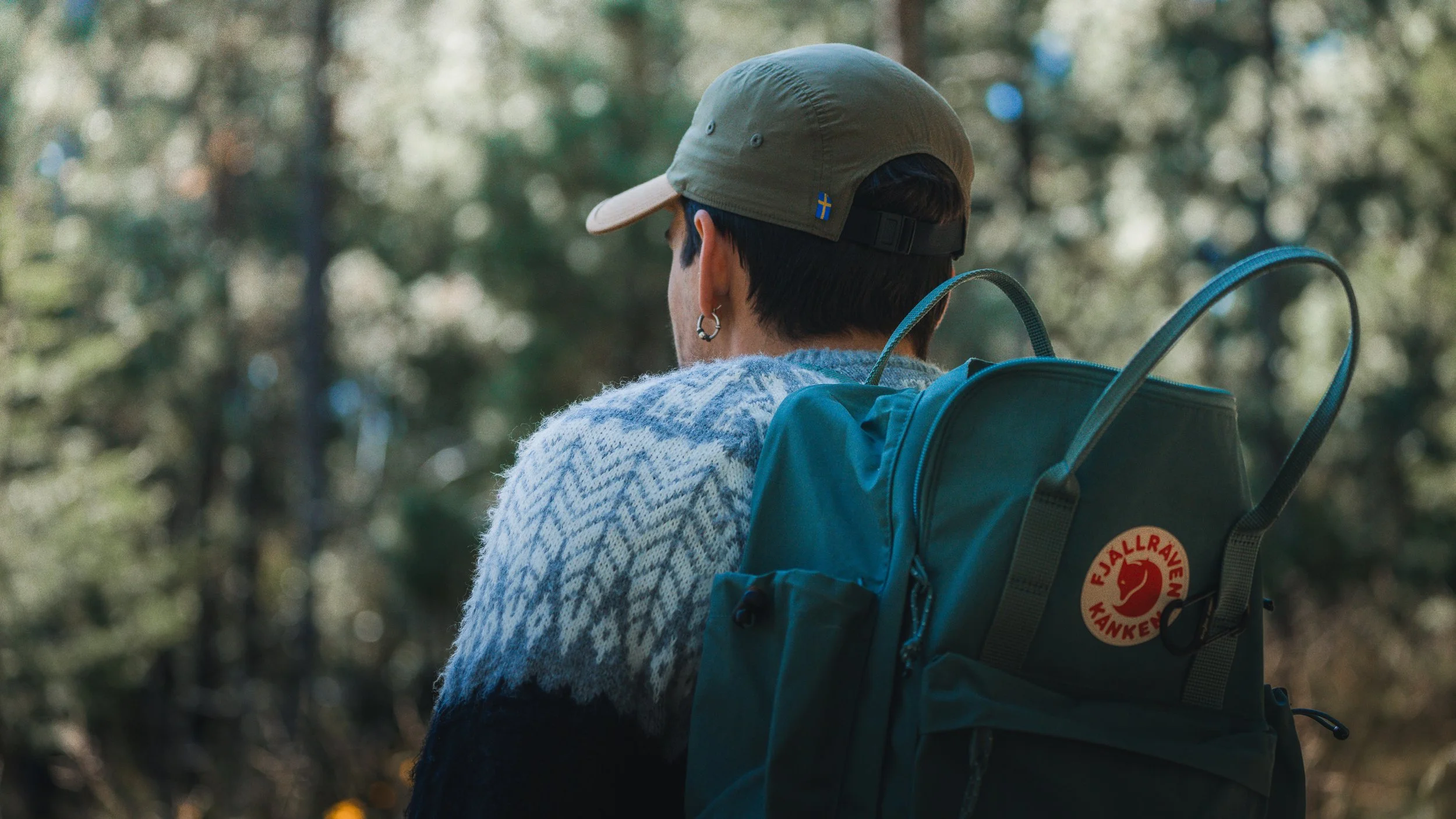 A person with short dark hair, wearing a beige cap with Swedish flag and earrings, looking away with a green Fjällräven backpack and a white patterned sweater, standing outdoors among trees.