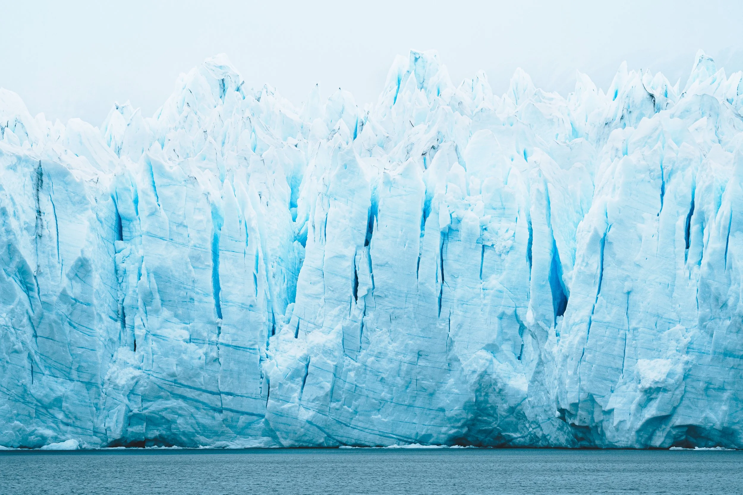 A large glacier with vertical ice formations, melting into a body of water in the foreground.