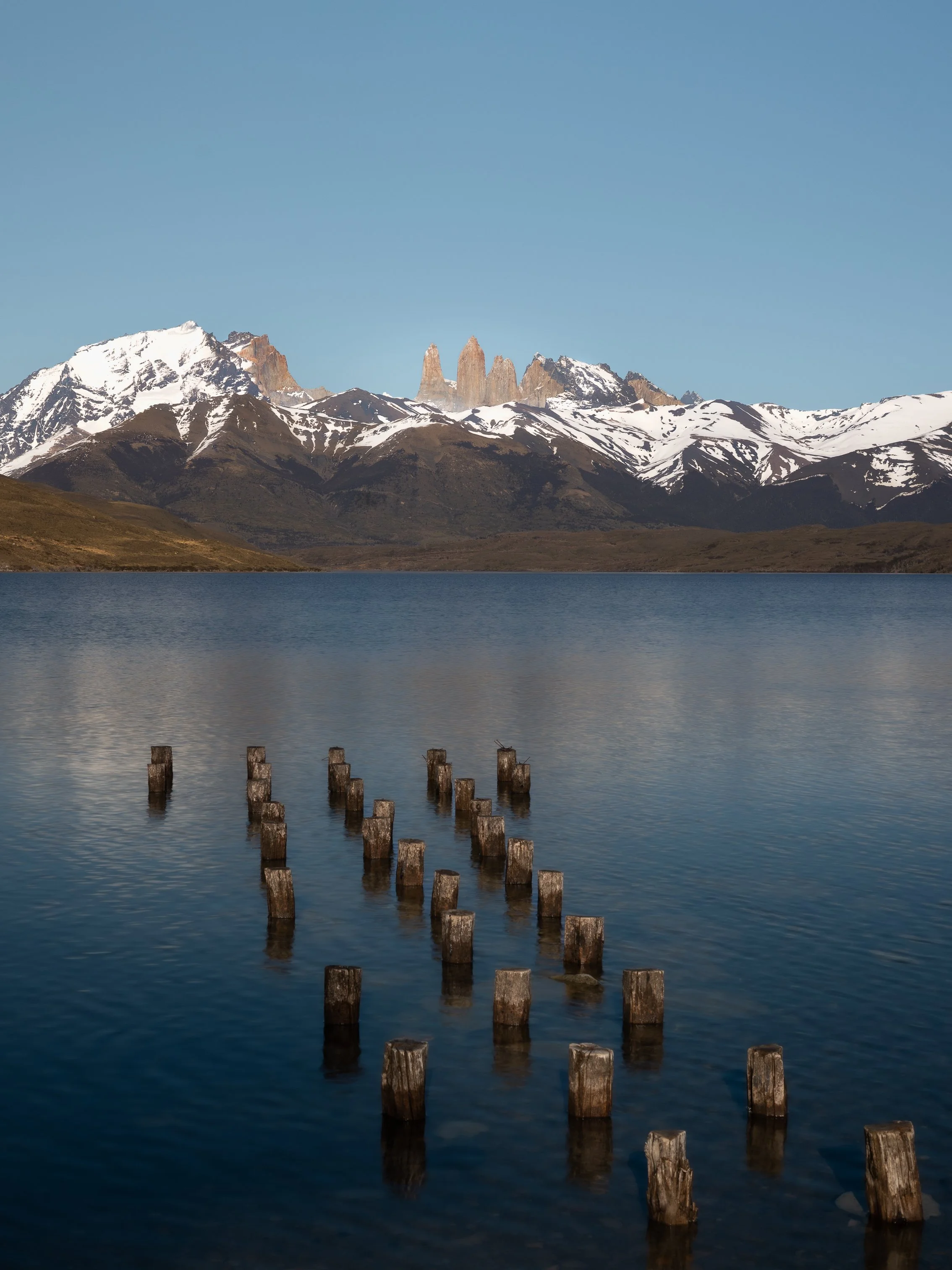 A mountain landscape with snow-capped peaks, a clear blue sky, and remnants of wooden posts in a calm lake in the foreground.