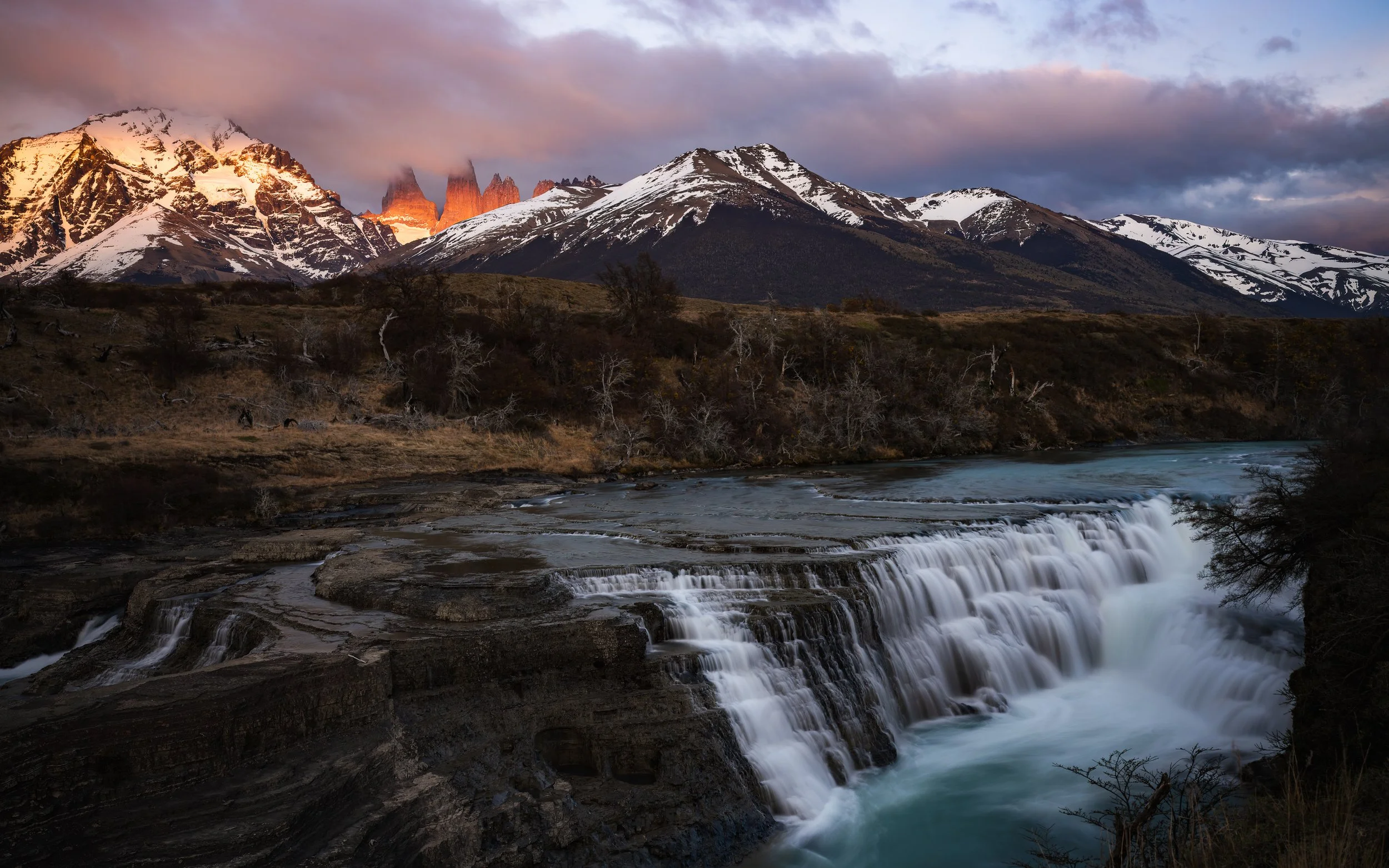 Scenic landscape featuring Torres del Paine a cascading waterfall in the foreground, dry trees along the riverbank, rugged mountains with snow-capped peaks in the background, and a partly cloudy sky at sunset with pink and purple hues.