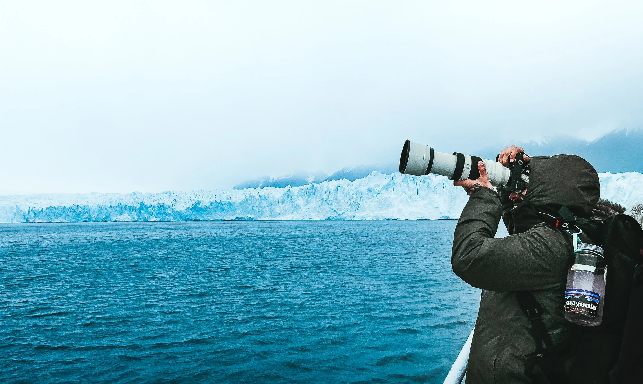 A person in a dark jacket and hood using a white telescope with a black eyepiece to observe icy mountains and glaciers across a body of water, on a cloudy day.