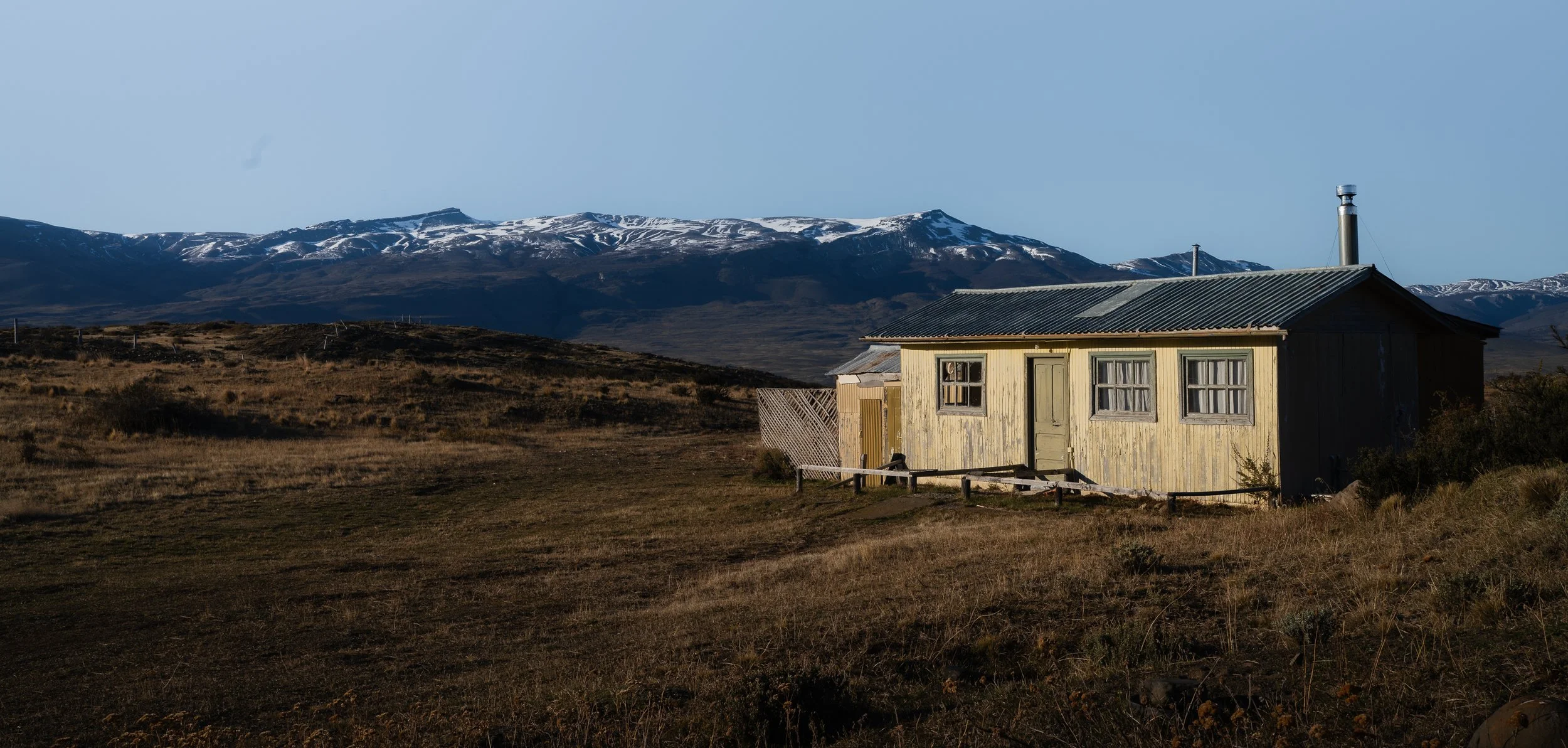A small, weathered yellow house with a tin roof sits in a barren landscape with mountains in the background.