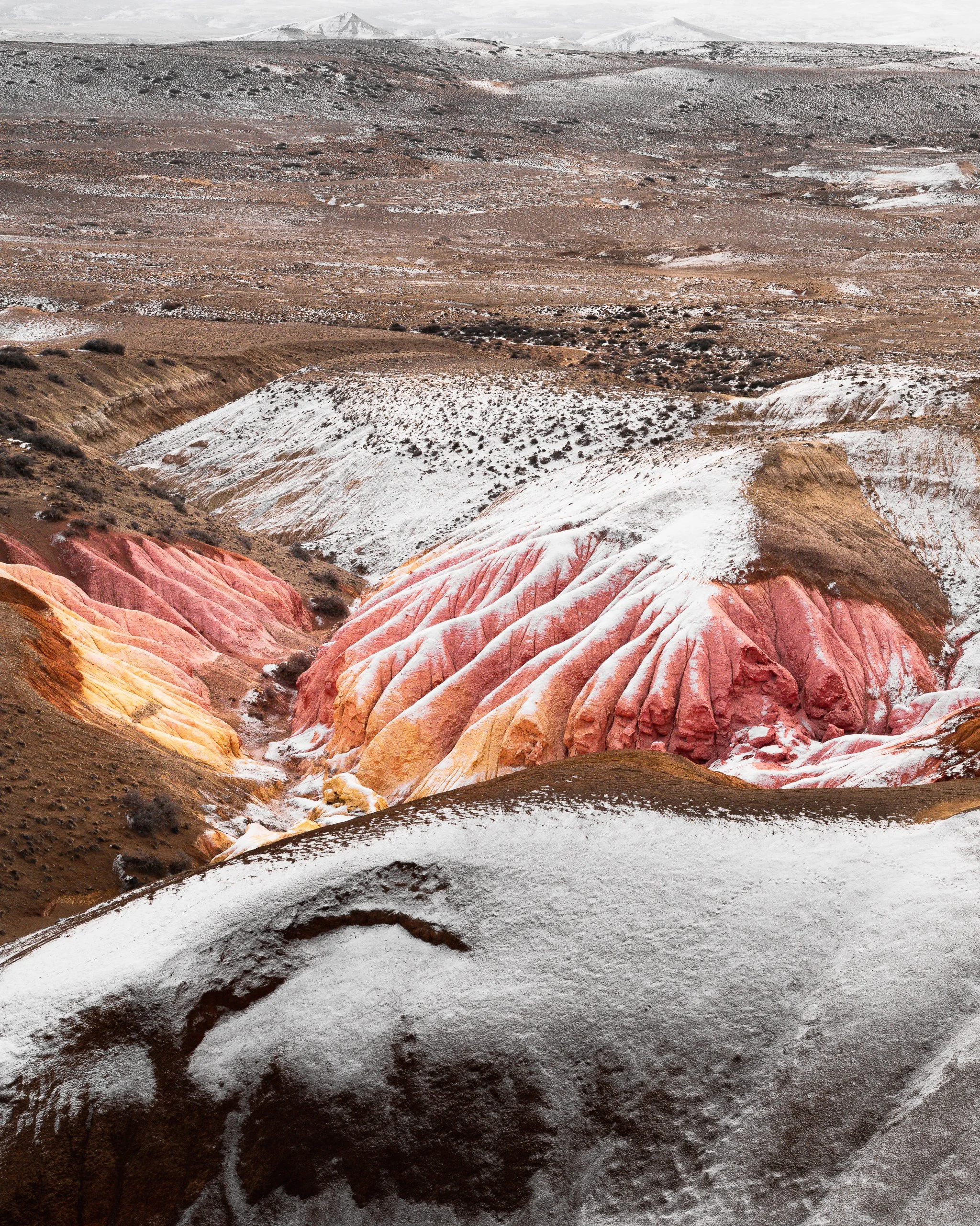 Colorful rock formation with red, orange, yellow, and white bands in a barren, snow-covered landscape with distant mountains.