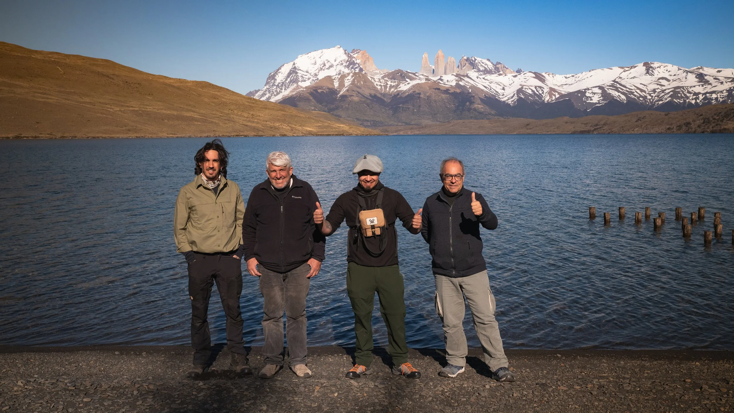Four men standing by a lake with snow-capped mountains in the background, smiling and giving thumbs-up.