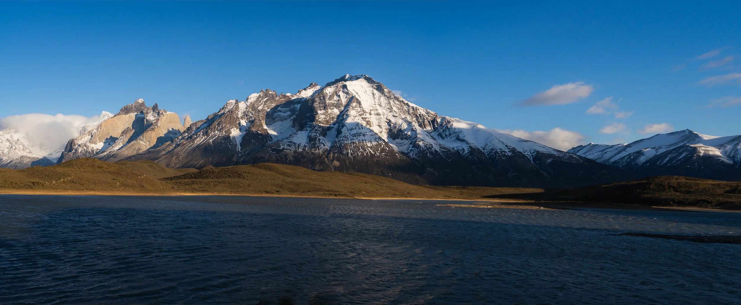 Snow-capped mountains behind a body of water with grassy hills in the foreground.