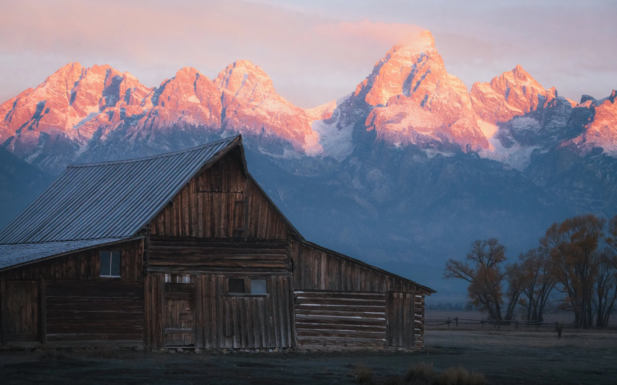 A rustic wooden barn with a metal roof, set against a backdrop of snow-capped mountains at sunrise or sunset, with some trees and a fence in the foreground in Grand Teton National Park
