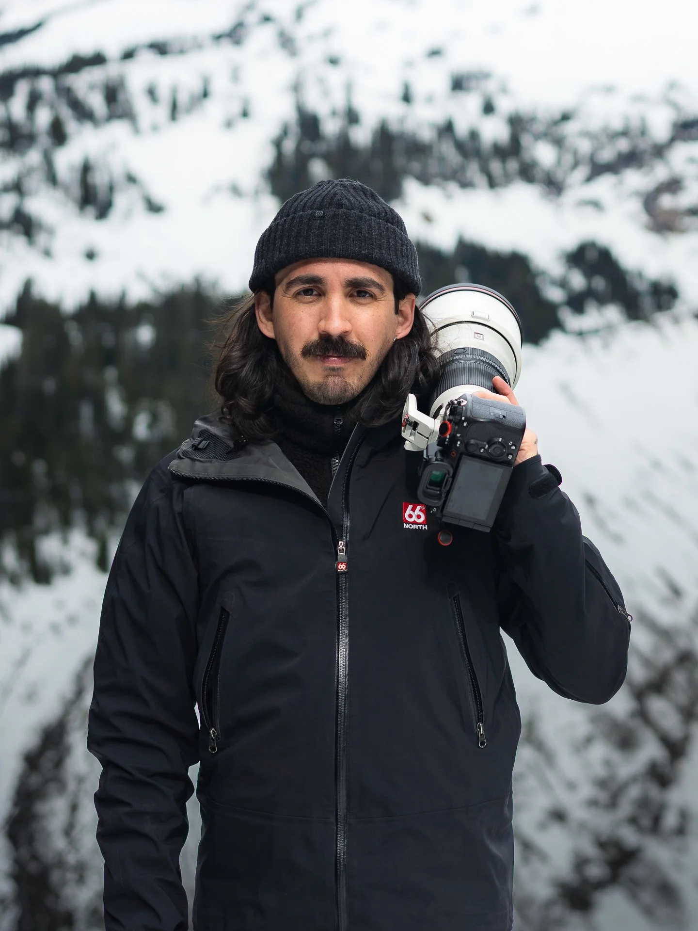 Fer Richaud: Wildlife Photographer: A man with long dark hair, a mustache, wearing a black beanie and black jacket, holding a camera with a large telephoto lens outdoors with snow-covered mountains and trees in the background.