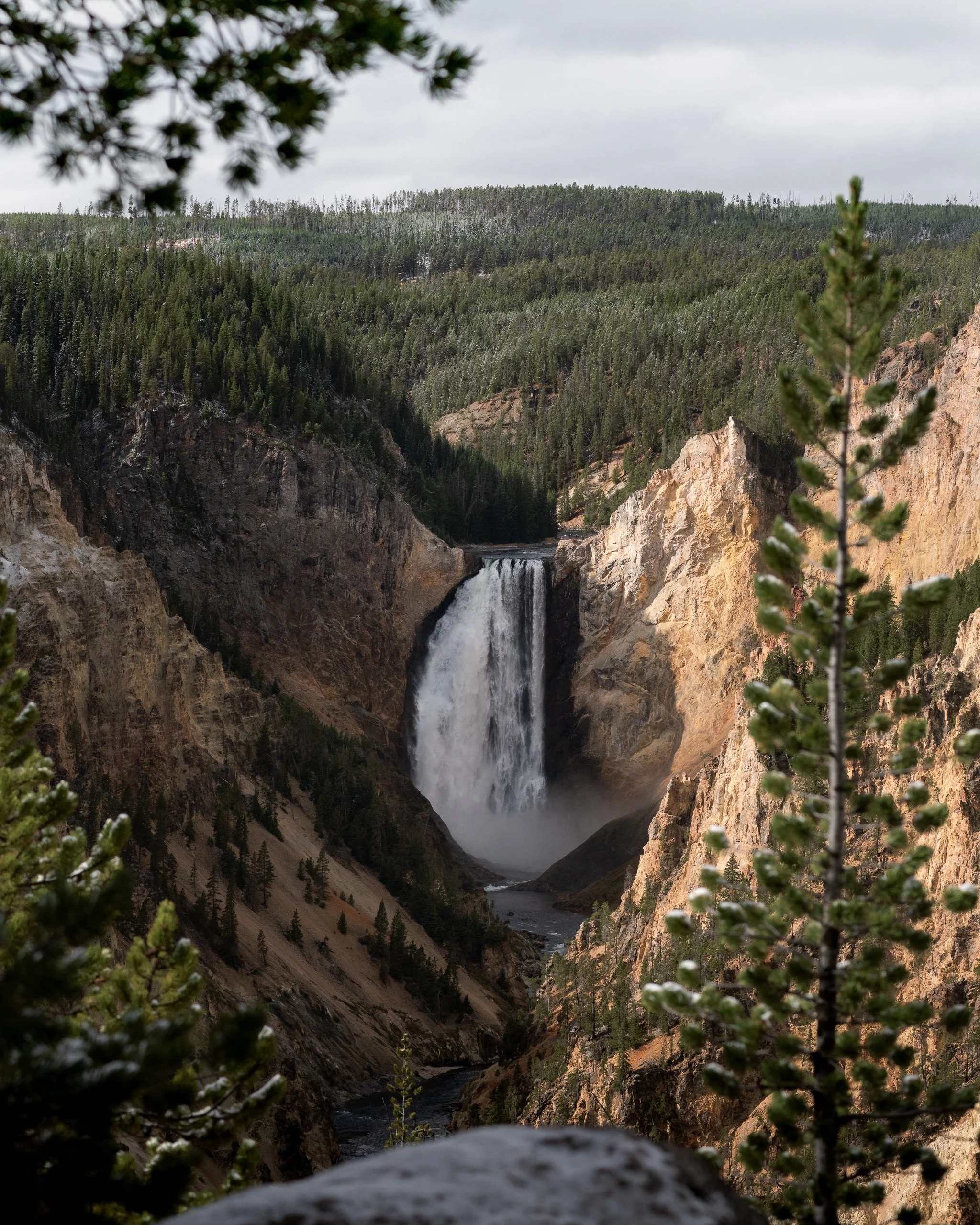 Waterfall in a canyon surrounded by forested mountains with trees and cloudy sky.
