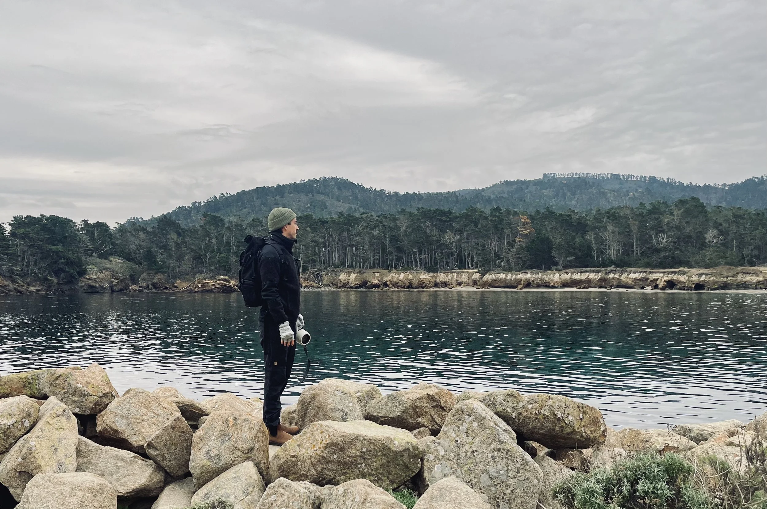 A man standing on rocks by a body of water, holding a rope with a loop at the end, with forested hills and an overcast sky in the background.