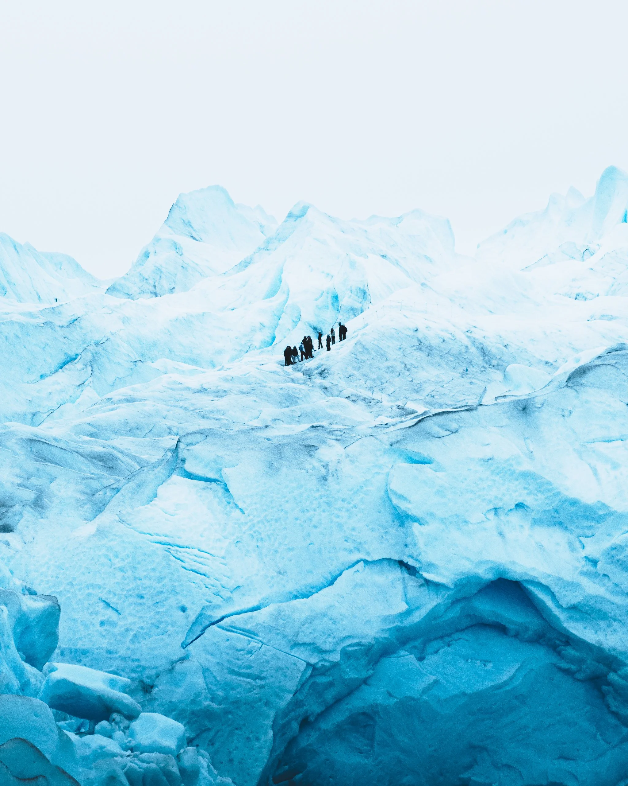 Group of people hiking on a glacier with ice formations and snow-covered peaks in the background.