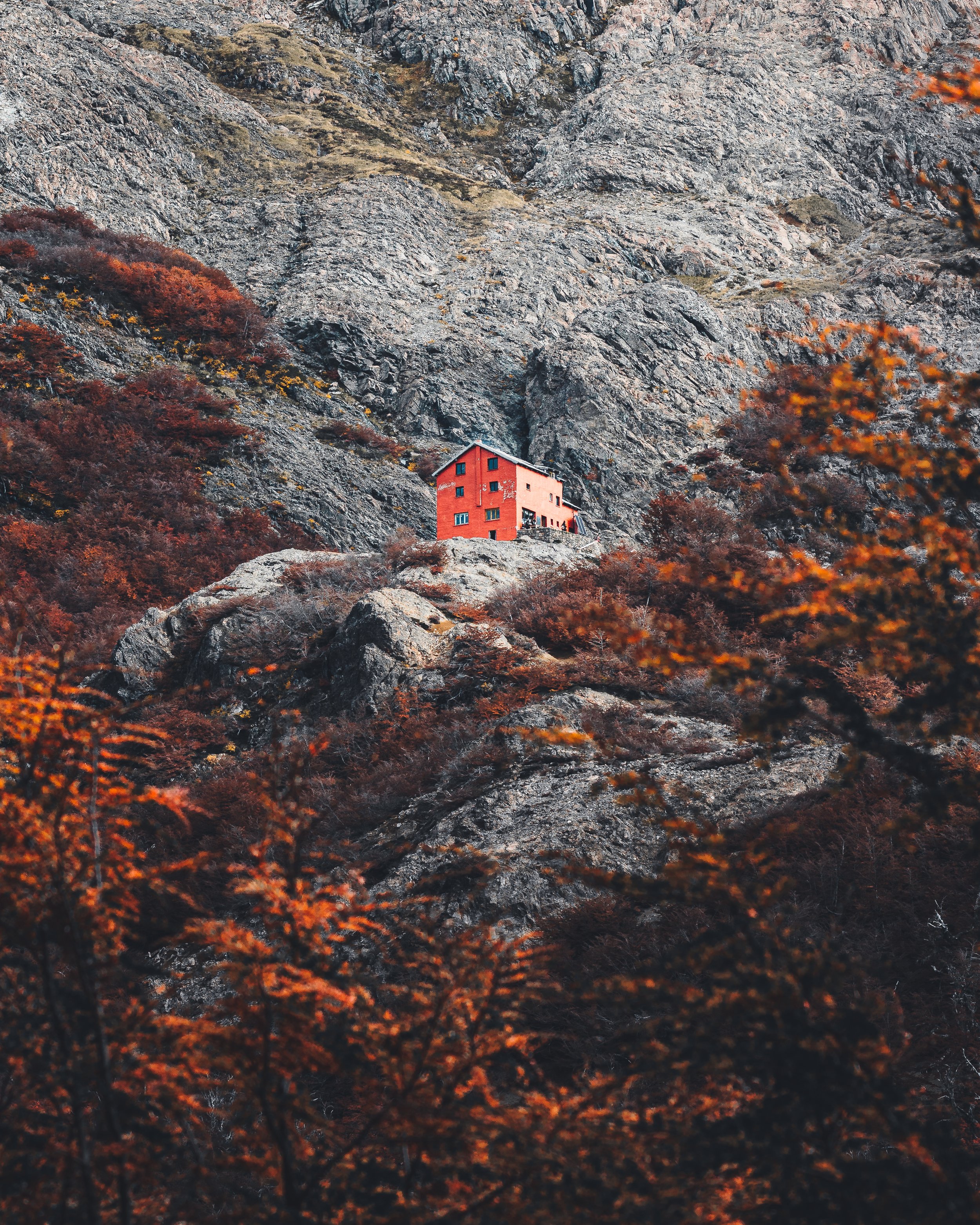 A red house perched on a rocky mountainside, surrounded by fall foliage with orange and brown leaves.