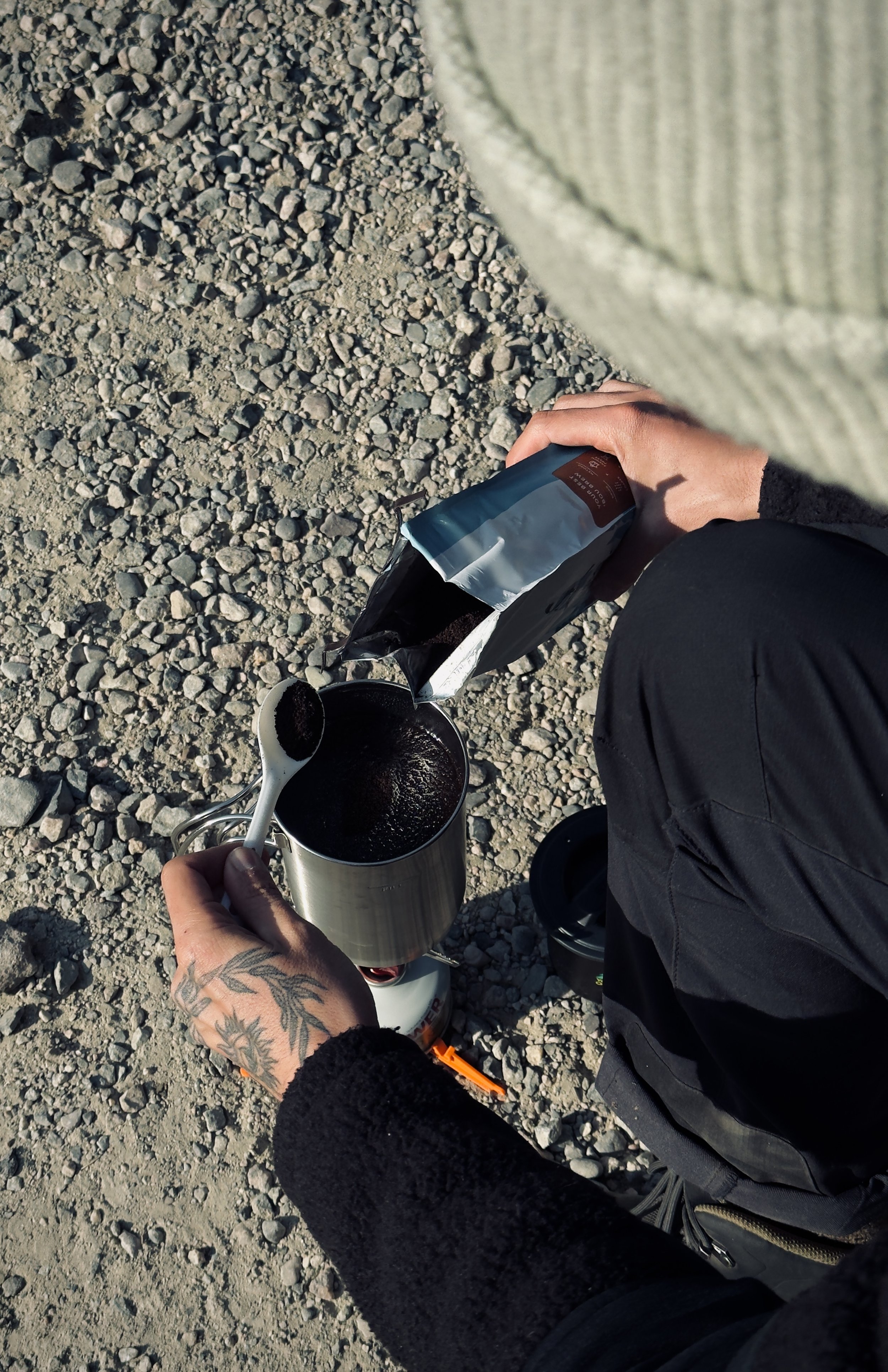 Person sits on rocky ground, preparing to pour coffee from a portable stove using a bag of coffee and a spoon.