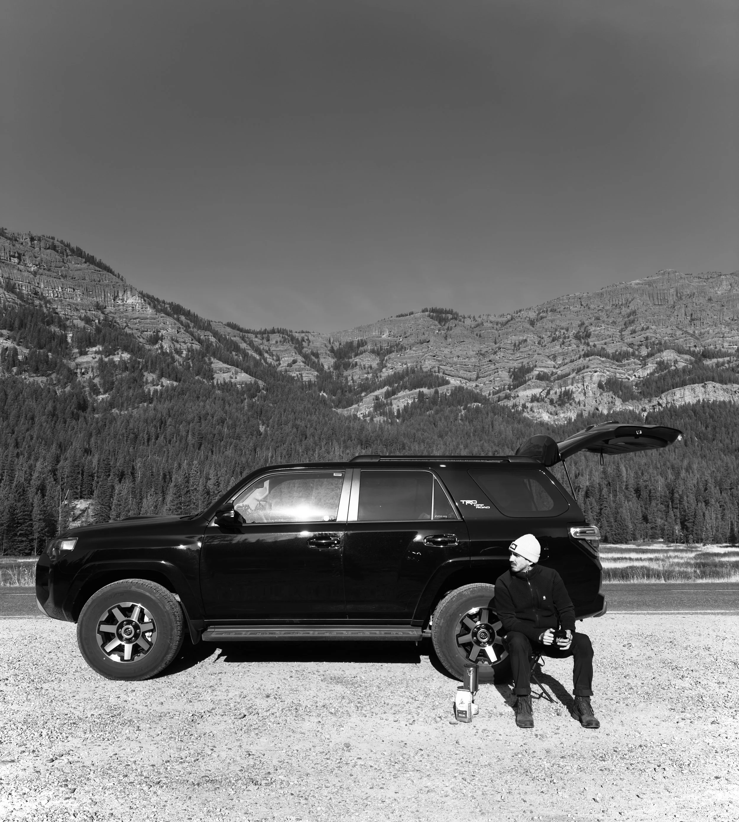 A man sitting on a small stool beside a parked black SUV in an open mountainous landscape, with mountains and trees in the background.