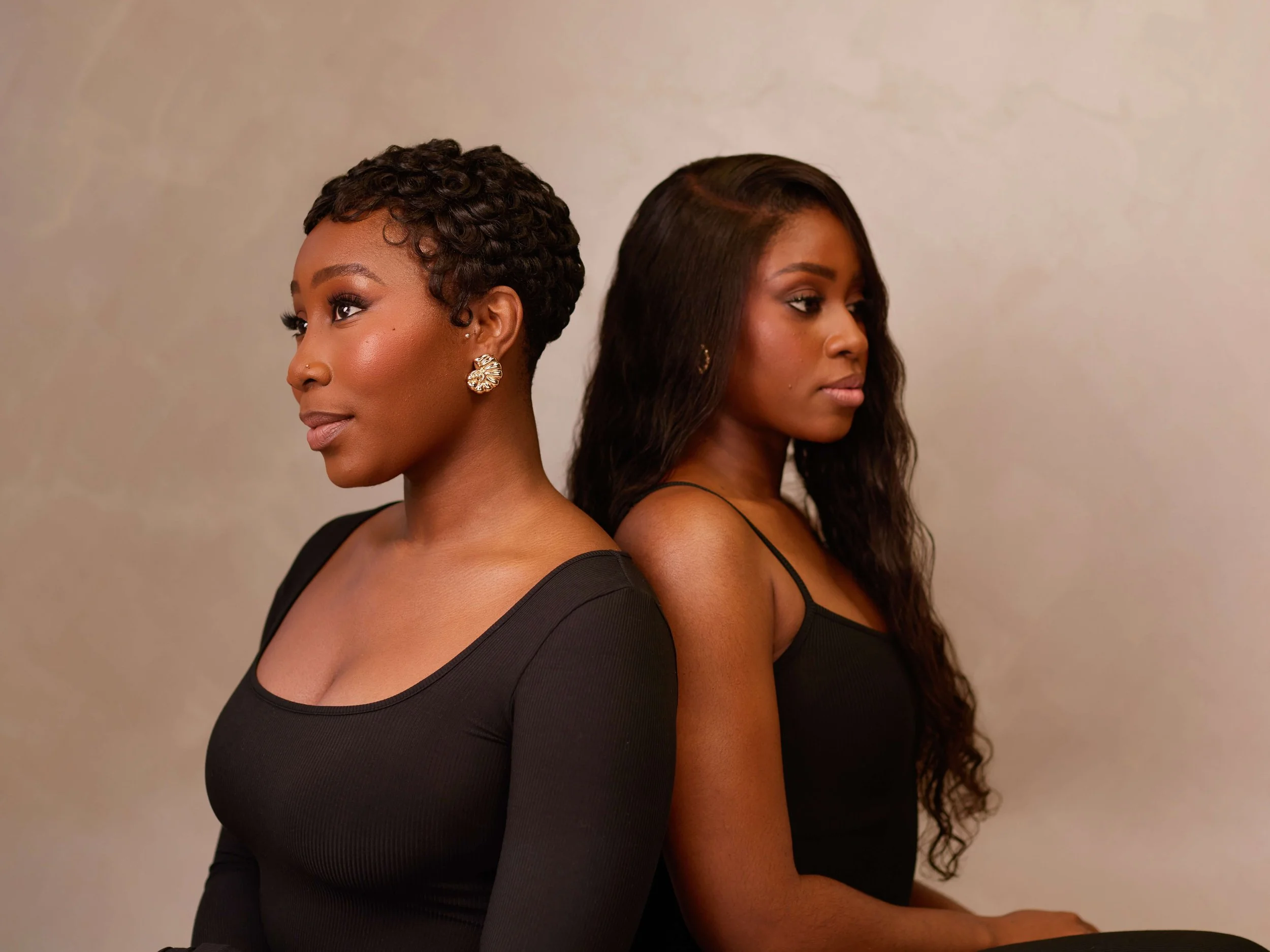 Two women with dark skin standing back-to-back against a plain wall, looking in opposite directions. The woman on the left has short, curly hair and wears a black top with gold earrings. The woman on the right has long, wavy hair and wears a black sleeveless top.