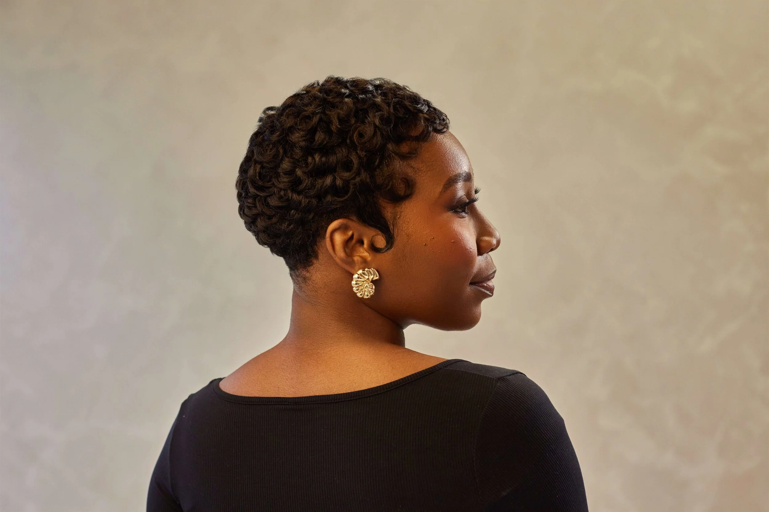 Side profile of an African American woman with short, curly hairstyle, gold earrings, wearing a black top, against a neutral background.