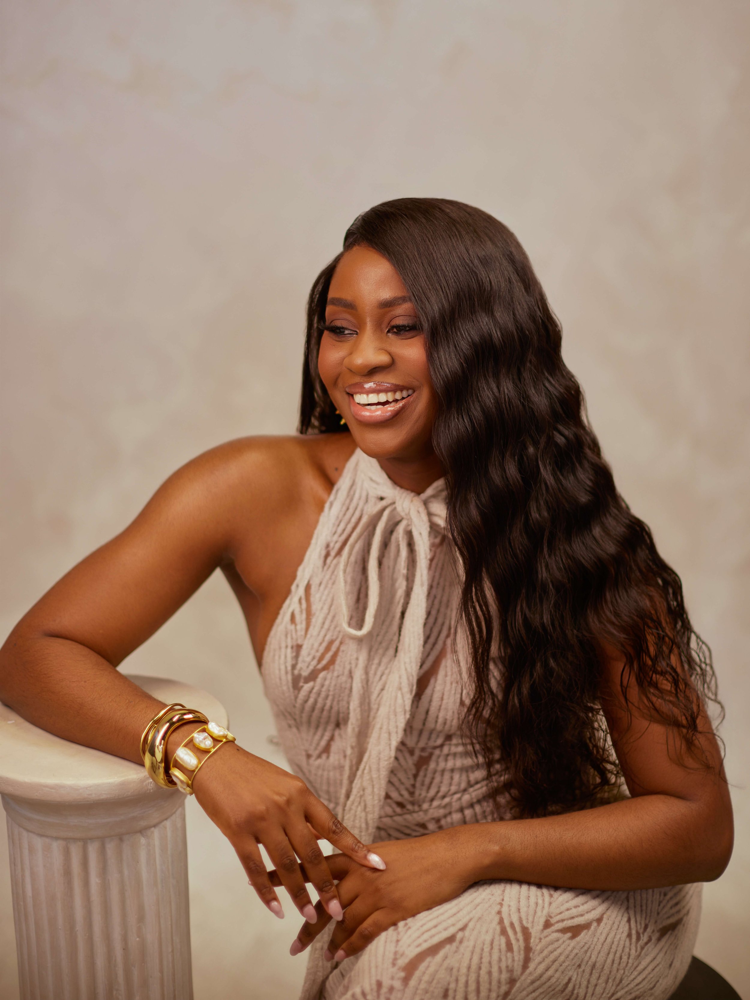 A woman with long wavy dark hair smiling, wearing a beige dress with a bow at the neck, gold bracelets, and sitting next to a white classical-style column, against a neutral background.