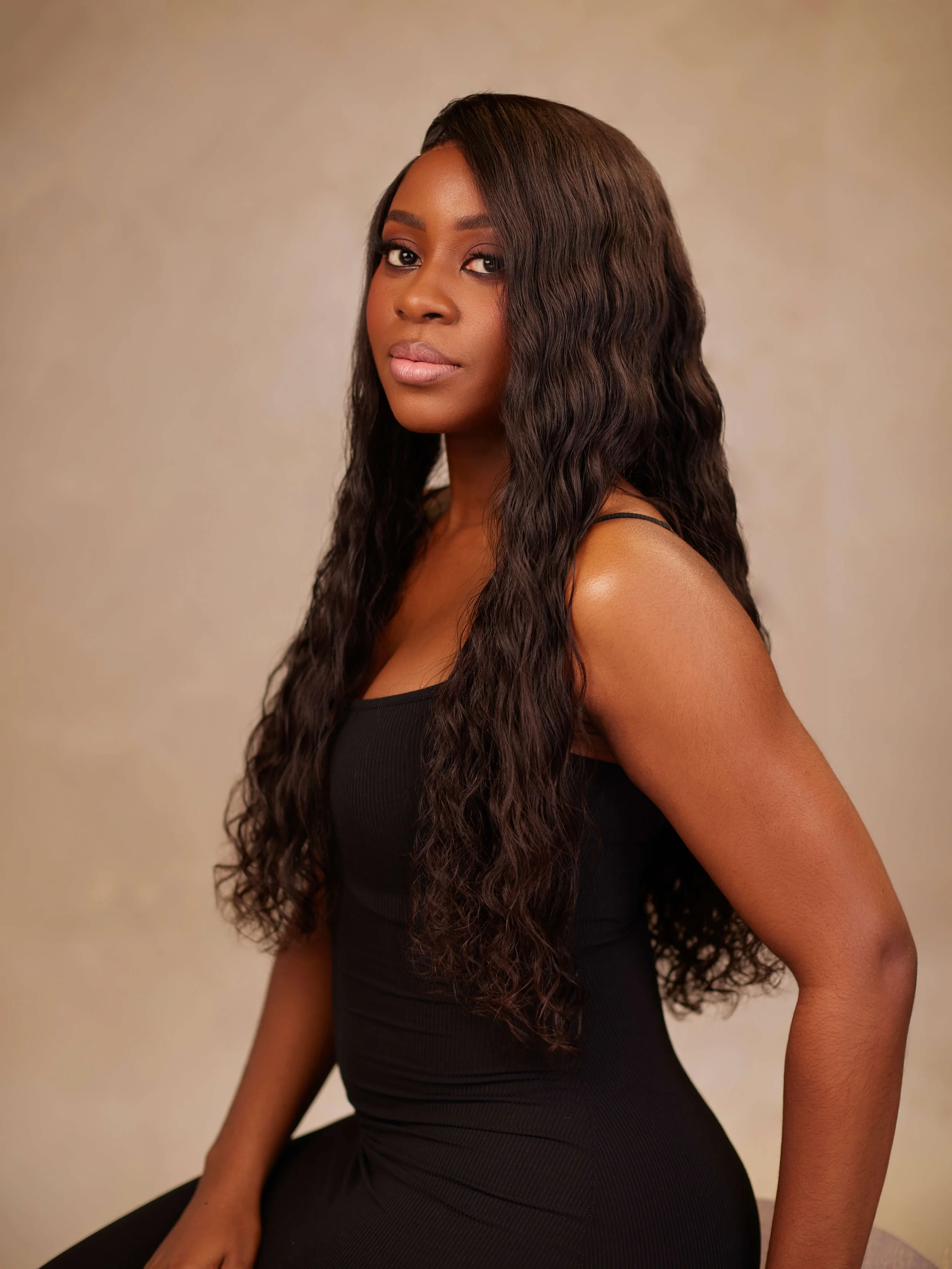 A young woman with long, wavy dark hair and a confident expression, wearing a fitted black sleeveless dress against a neutral background.