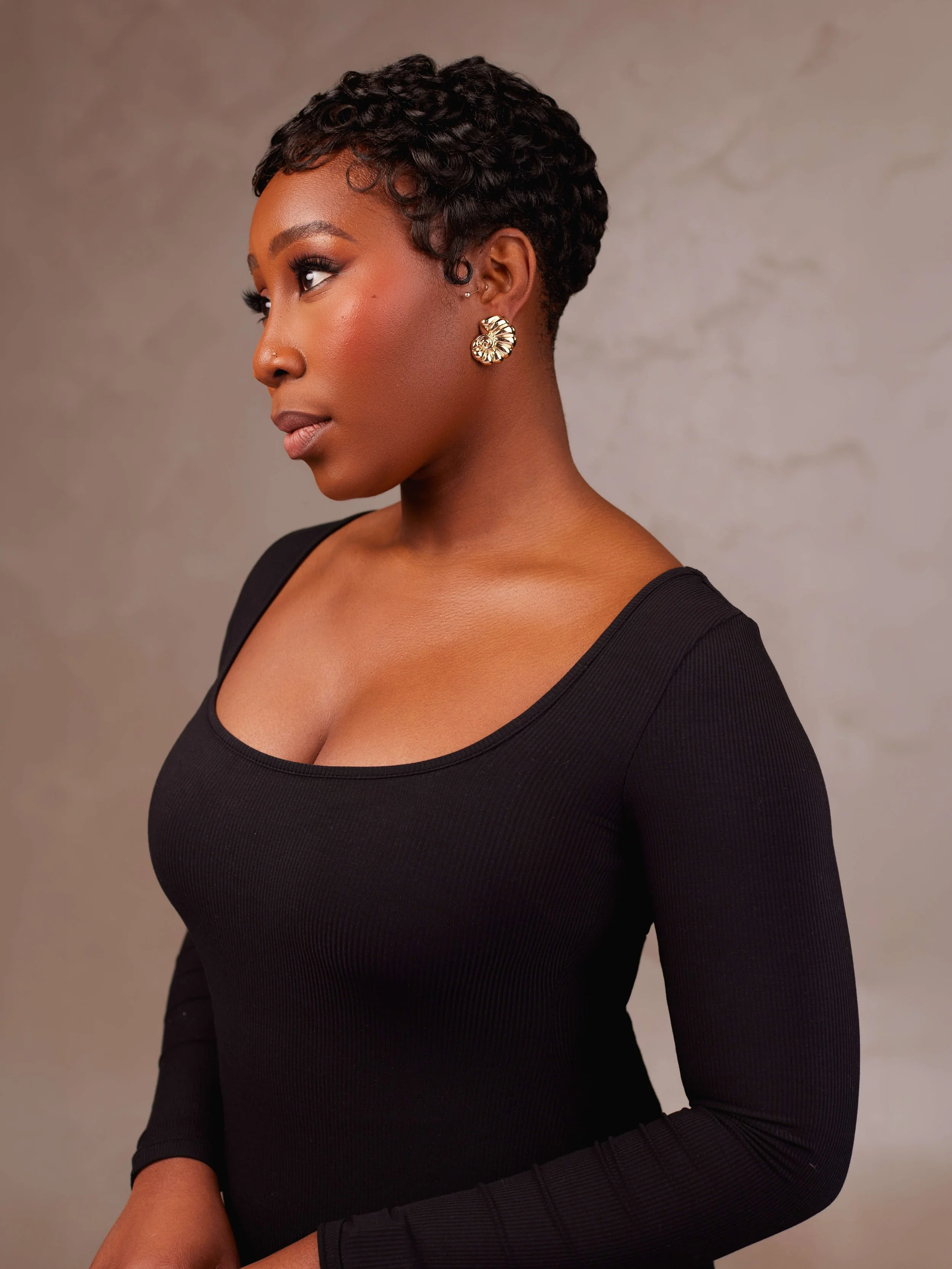 A woman with short, curly black hair wearing gold earrings and a black long-sleeve top, looking to her left against a neutral background.