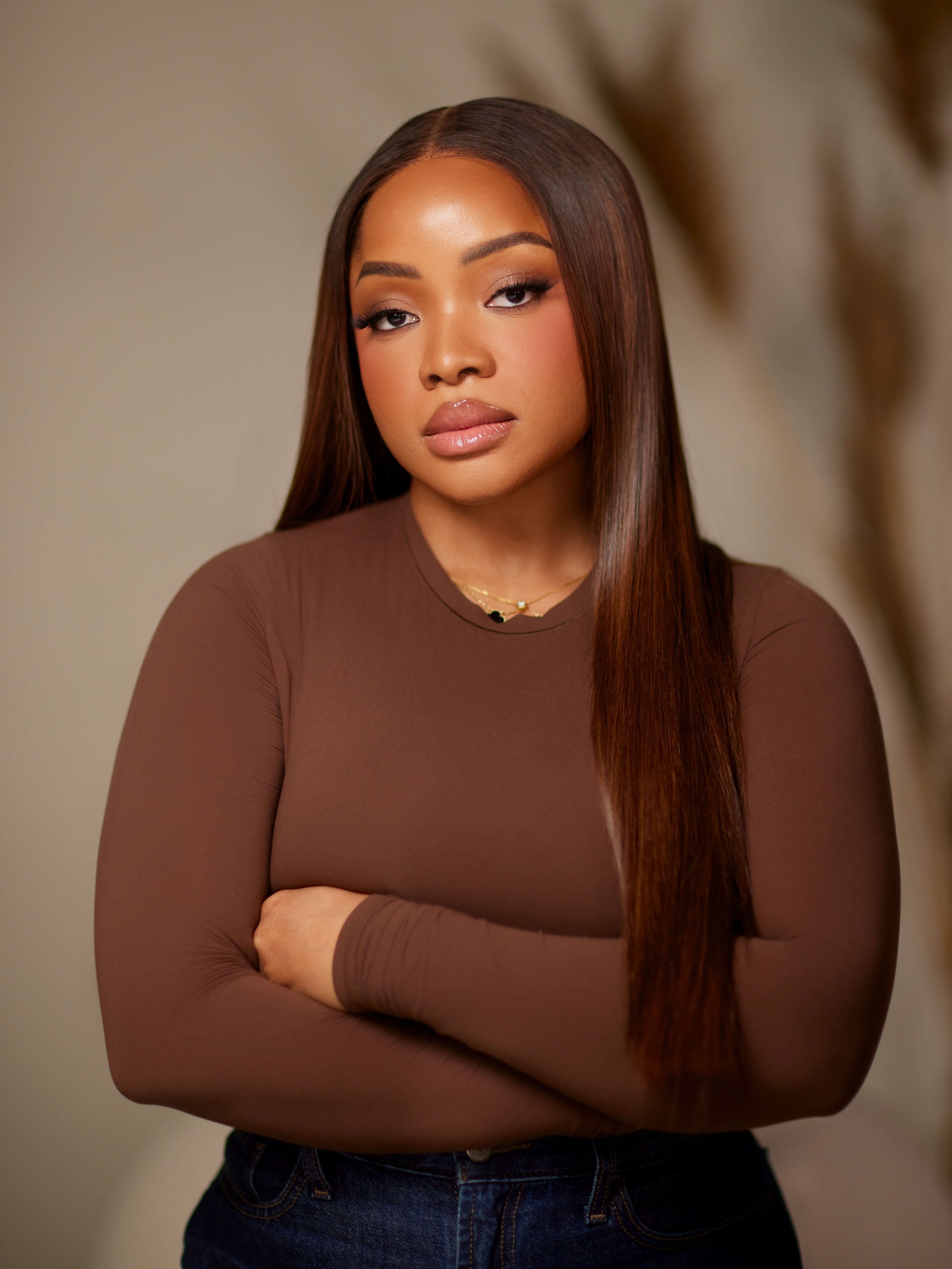 A woman with straight brown hair, wearing a brown long-sleeve top and blue jeans, standing with arms crossed in front of a neutral background.