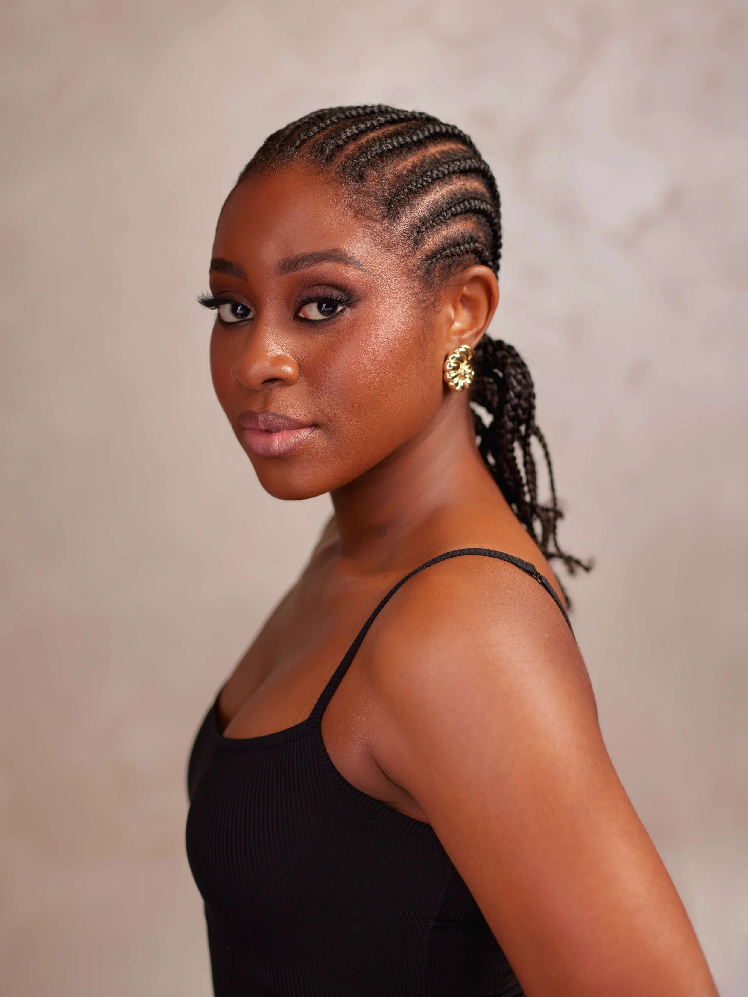 A young Black woman with braided hair wearing a black spaghetti strap top and gold earrings, posing against a neutral background.