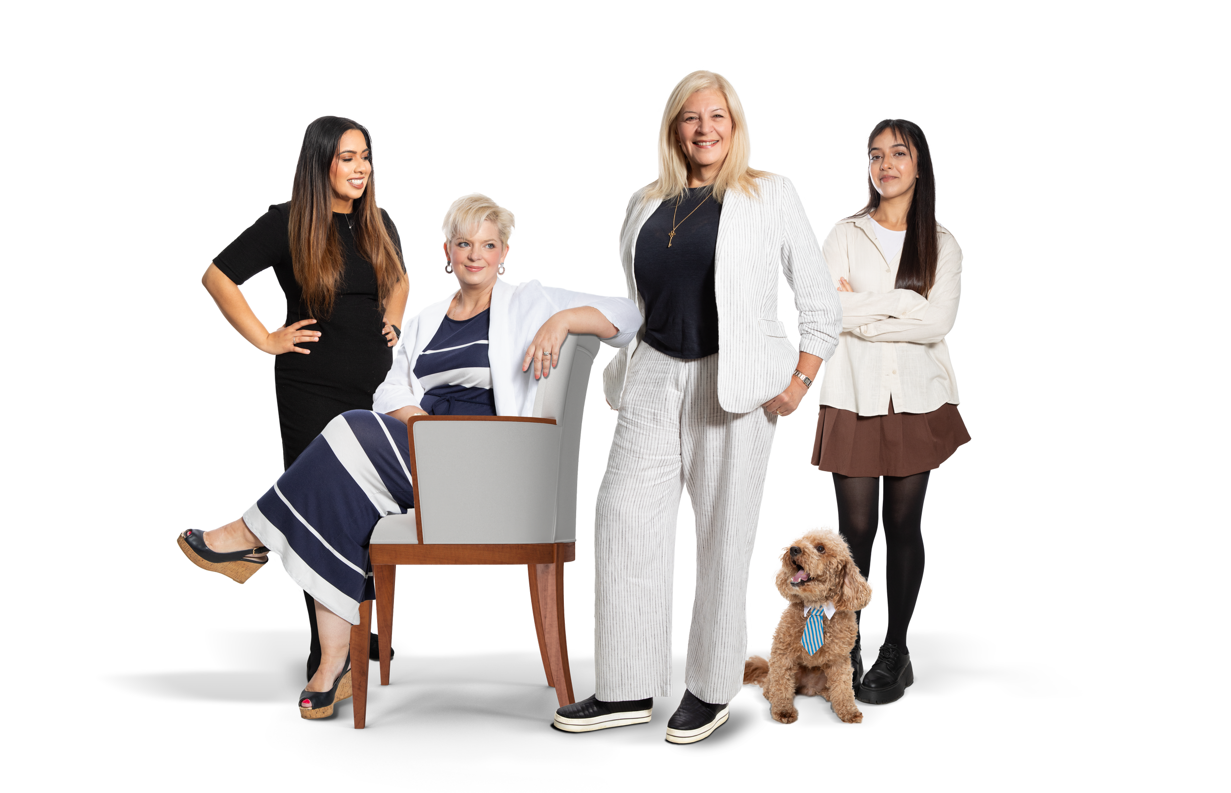 Group of five women and a dog posing together indoors. The women are diverse in age and style, with one sitting, three standing, and a dog sitting on the floor.