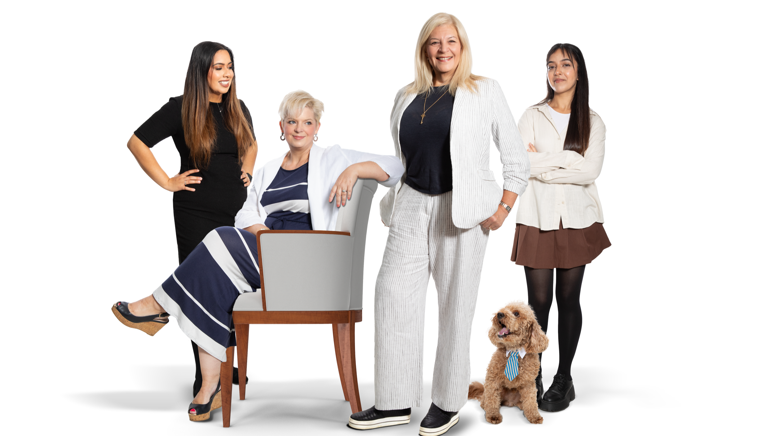 A group of four women and a dog posing together. One woman is sitting on a chair, wearing a striped dress. Two women are standing, one with crossed arms and the other with her hand in her pocket. The fourth woman is standing with her hands on her hips, and a golden doodle dog is sitting in front of them.
