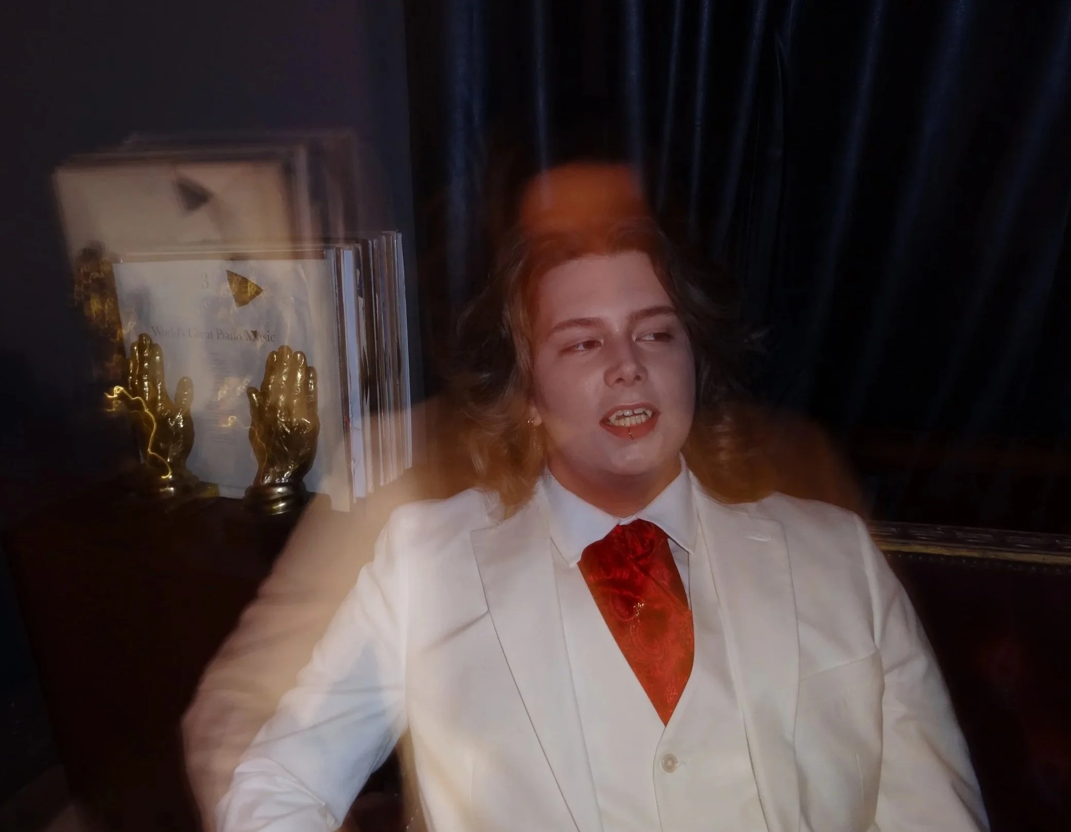 Person with long hair wearing white suit and red tie, sitting in front of a dark curtain, with blurred objects on the left side.