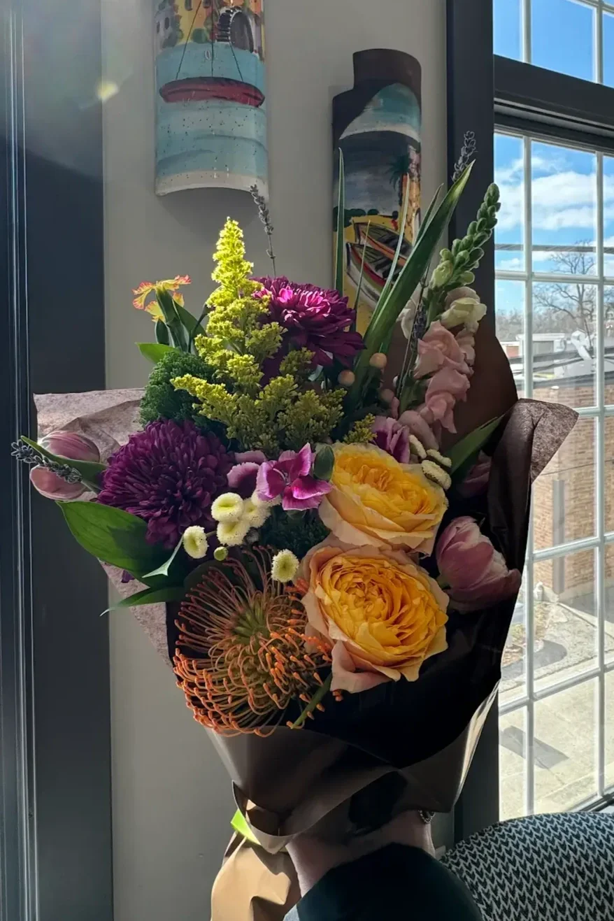 A lush, colorful floral bouquet wrapped in dark brown paper, featuring large golden-yellow garden roses, an orange pincushion protea, deep purple chrysanthemums, and light pink tulips.