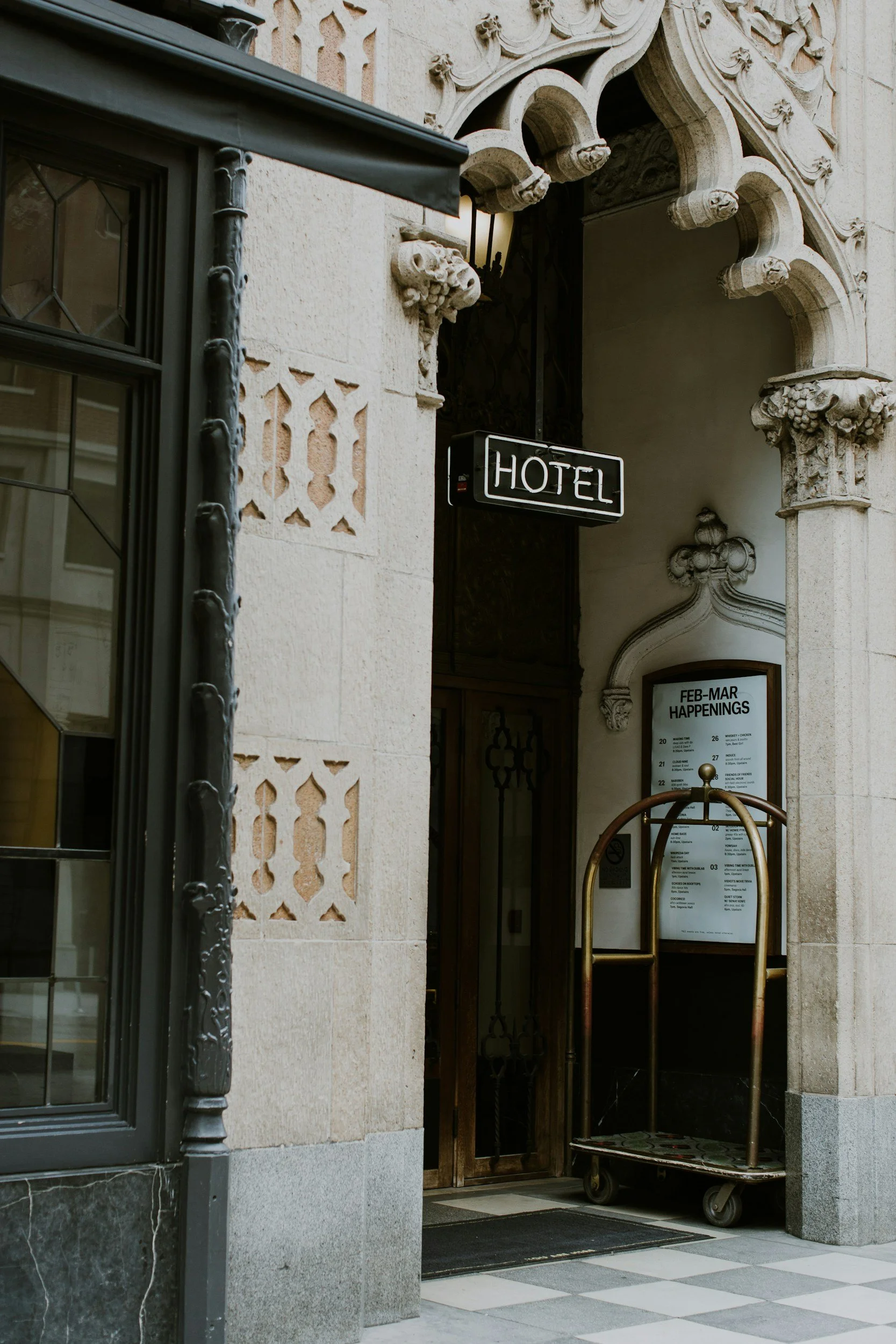 Hotel entrance with ornate architectural features, a black and white neon sign reading 'HOTEL', a luggage cart, and a board listing February-March events.