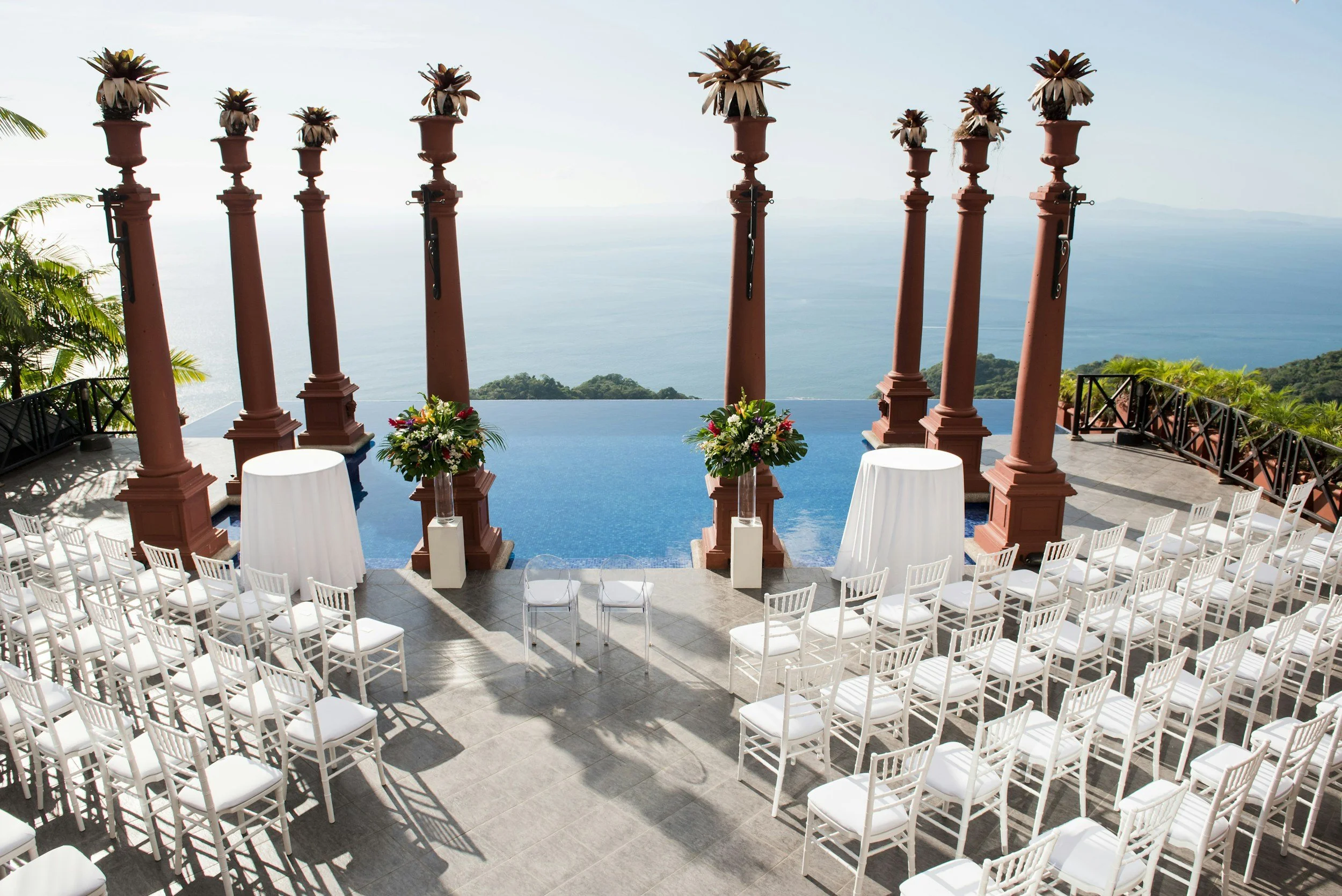 Outdoor wedding setup with white chairs, floral arrangements, and a view of the ocean and mountains in the background.