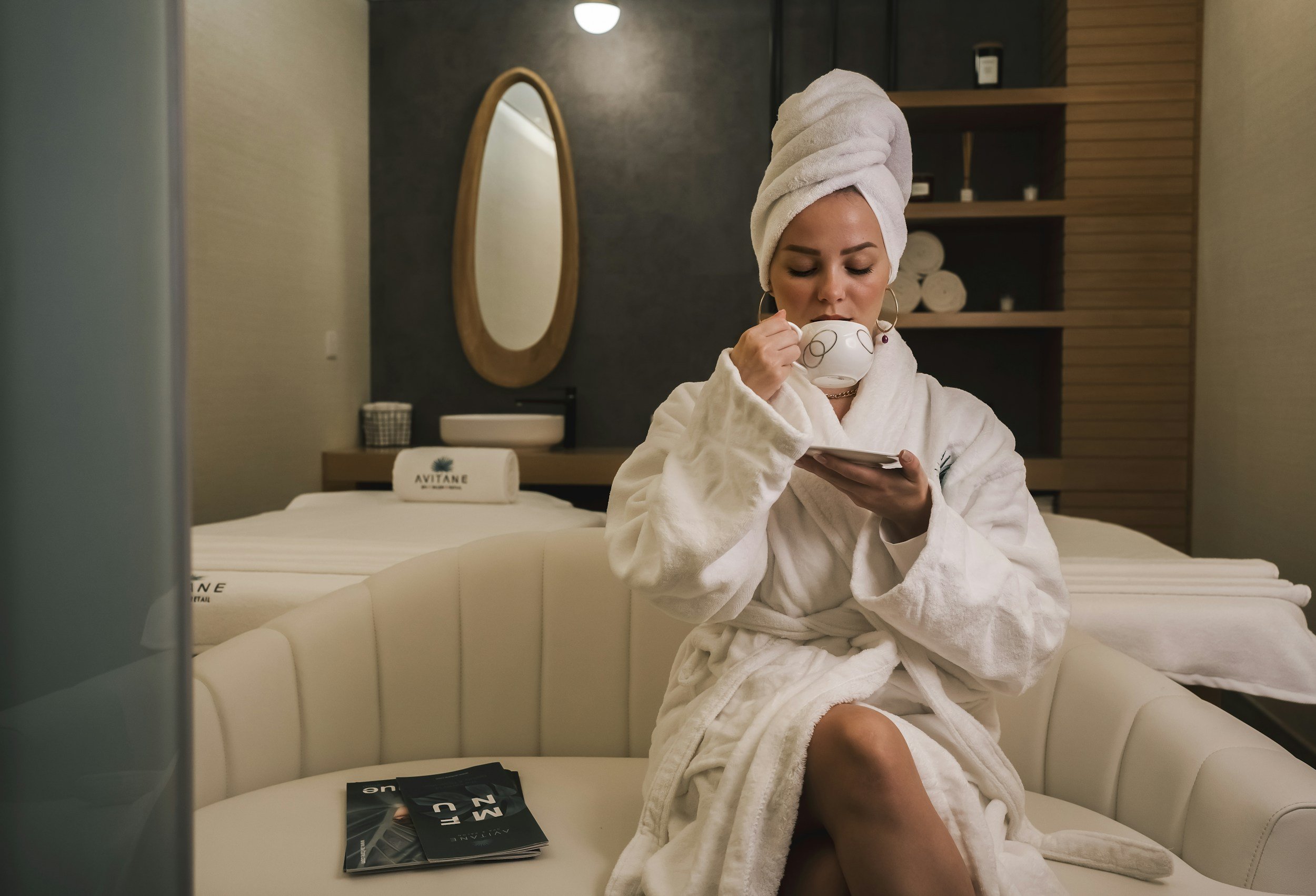 A woman in a white bathrobe and towel wrapped around her head, sitting on a cream sofa in a wellness or spa room, sipping from a cup. She is looking at her phone, with magazines on the sofa beside her and a bed with a towel and amenities in the background.