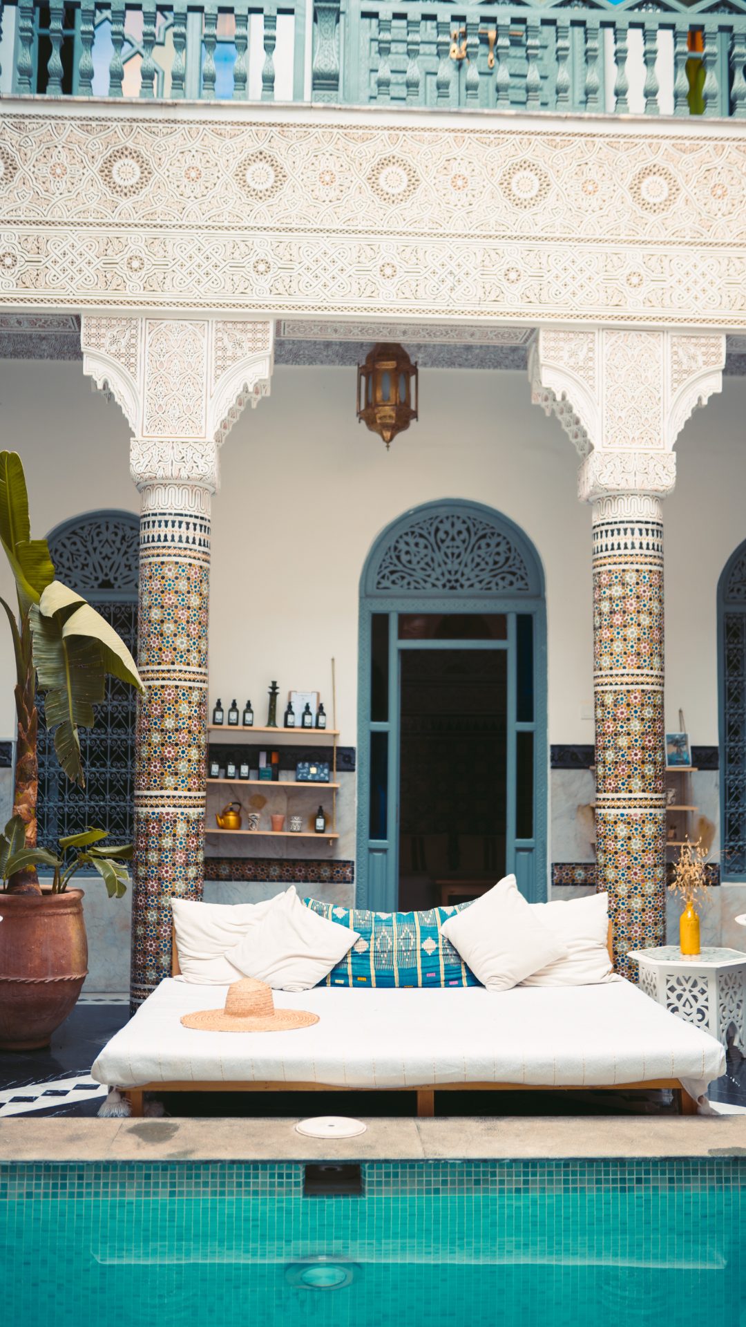 A Moroccan-style porch area with a pool in the foreground, decorative columns with colorful mosaic tiles, a sofa with pillows, a straw hat, and a potted plant, with blue door and windows, and intricate white architectural details.