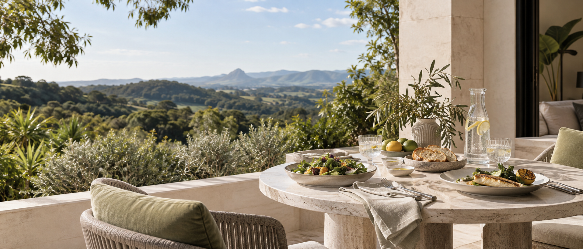 Outdoor dining table set with salads, bread, lemons, limes, water, and a pitcher, overlooking rolling hills and mountains in the distance on a sunny day.