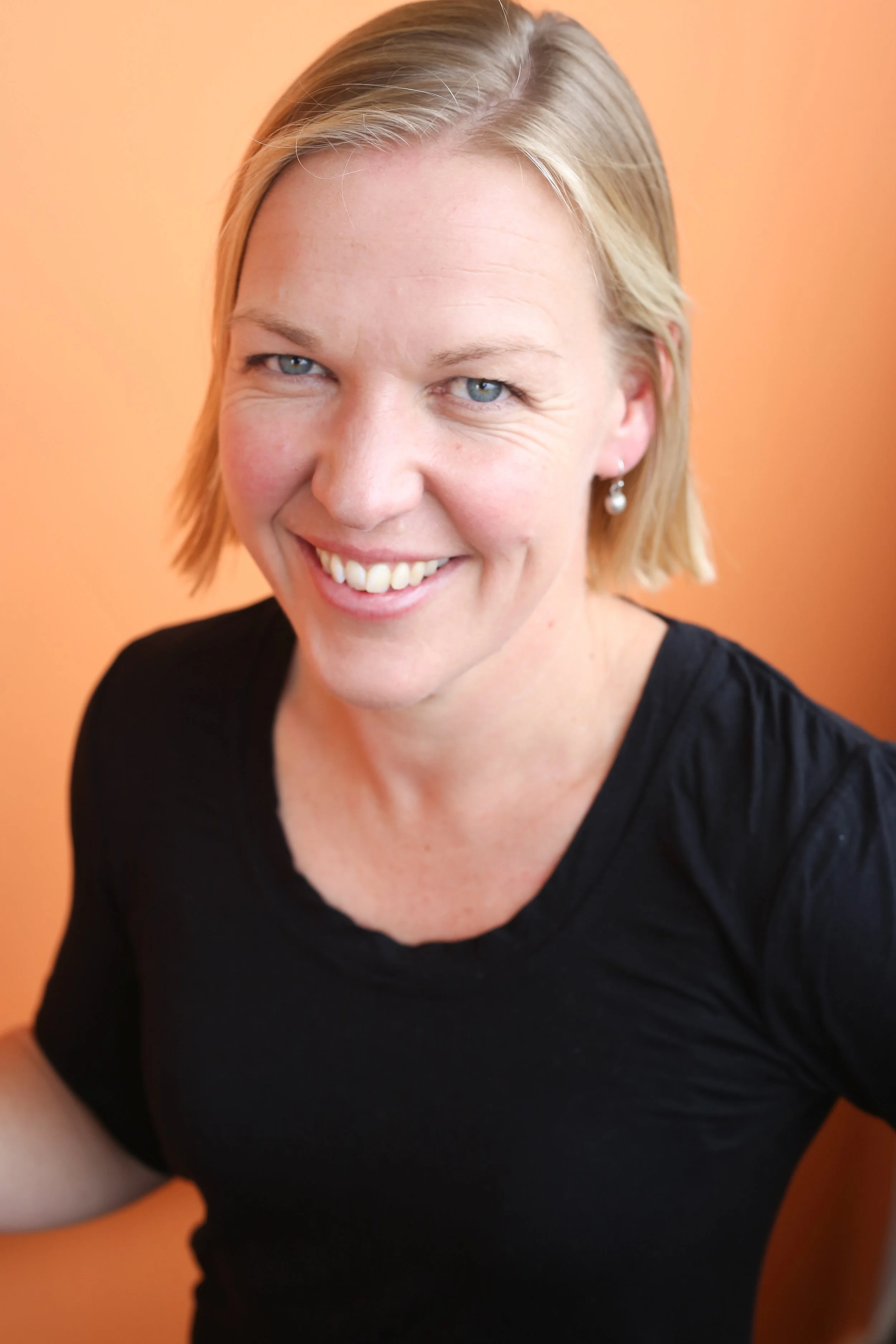 Close-up portrait of a woman with blonde hair wearing a black top and pearl earrings, smiling against an orange background.