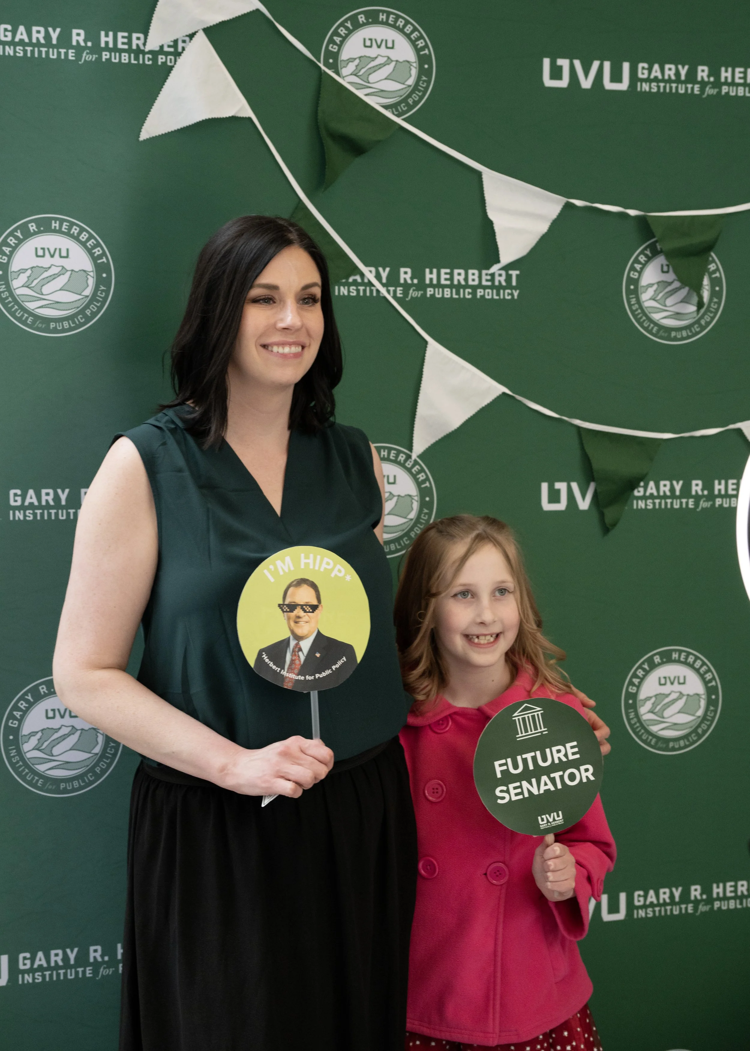 Two women, a young girl and her mom, standing in front of a green backdrop with logos of the Gary R. Herbert Institute for Public Policy. The women are holding circular signs; the young girl’s sign reads 'Future Senator'
