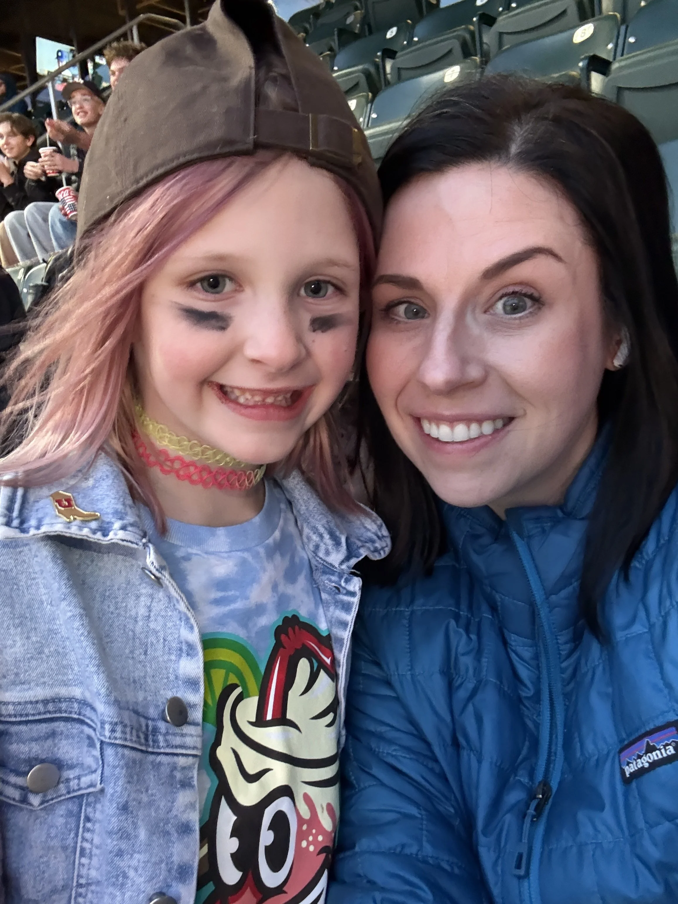 Two smiling women taking a selfie at a sports stadium with green seats and spectators in the background.
