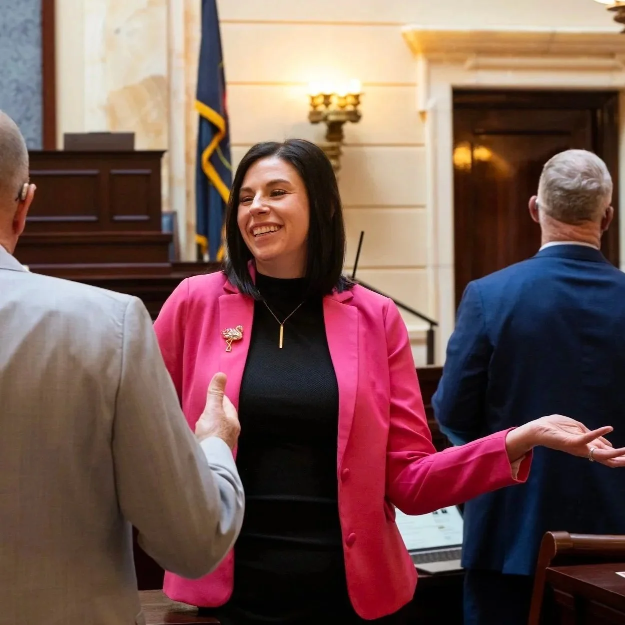 A woman in a pink blazer smiling and talking to two men in a formal setting with wooden paneling, a flag, and a pin on her blazer.