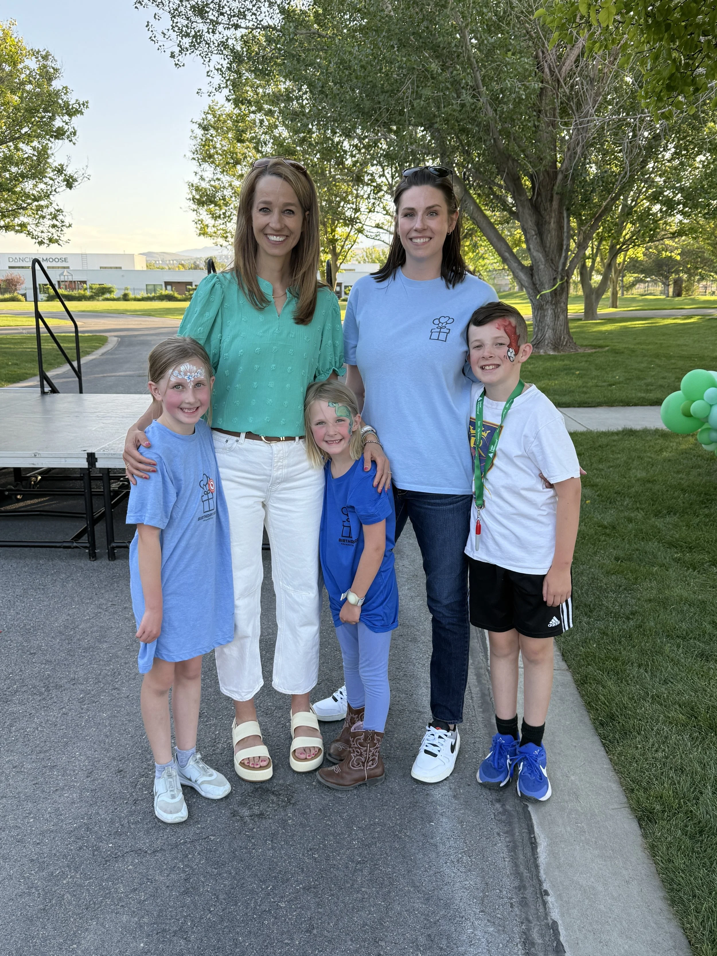 A group of five smiling people, including two women and three children, standing outdoors in a park with trees and a building in the background. The children have face paint.