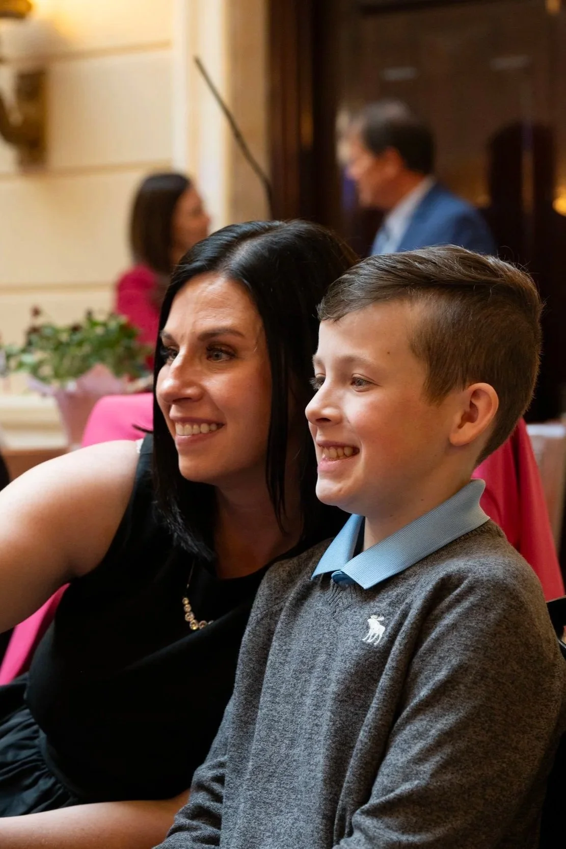 A woman and a boy sitting close together, smiling and taking a selfie at a social event or gathering, with other people blurred in the background.