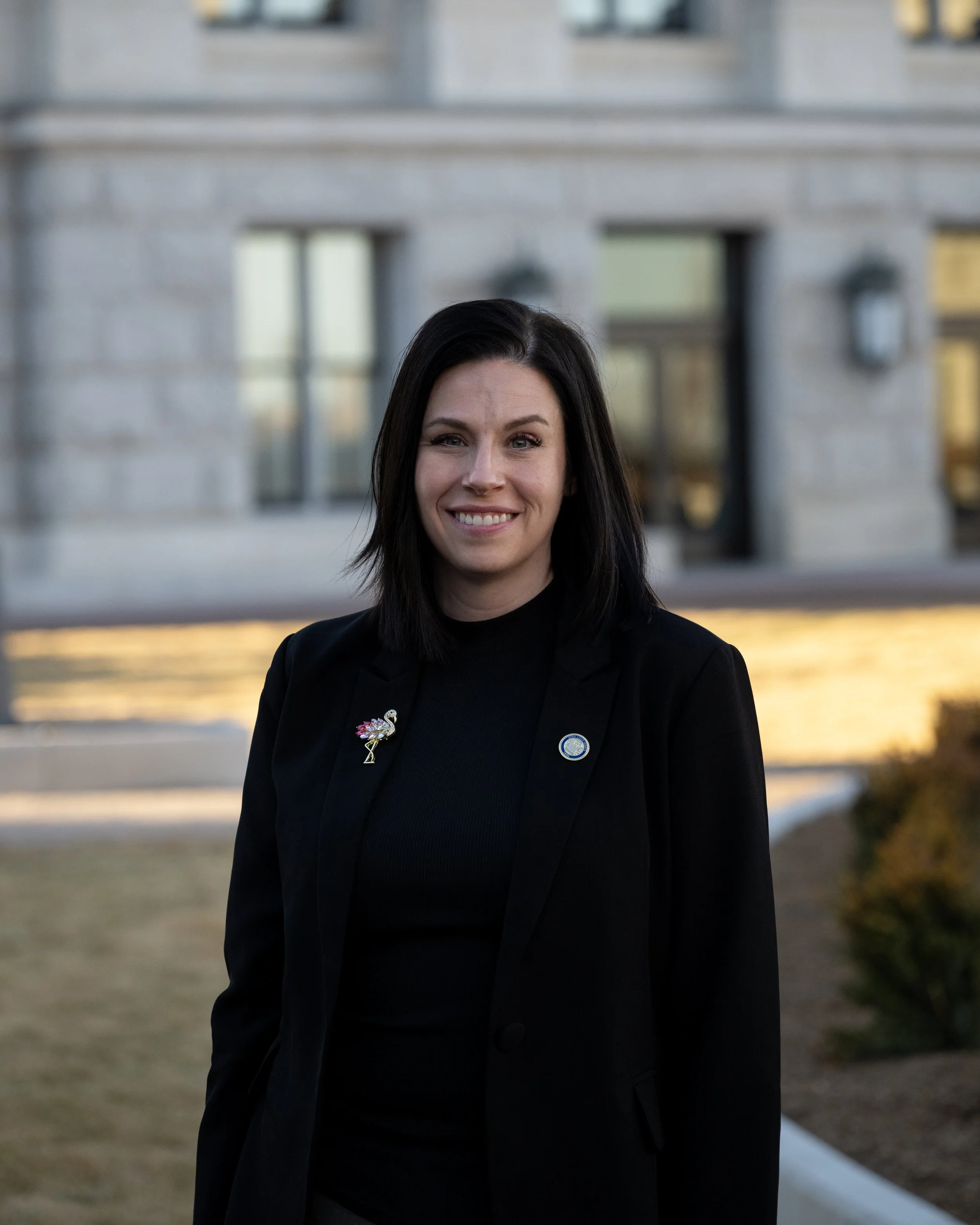 A woman with dark hair smiling, wearing a black blazer with pins, standing outdoors in front of a building with blurred windows and a water feature.