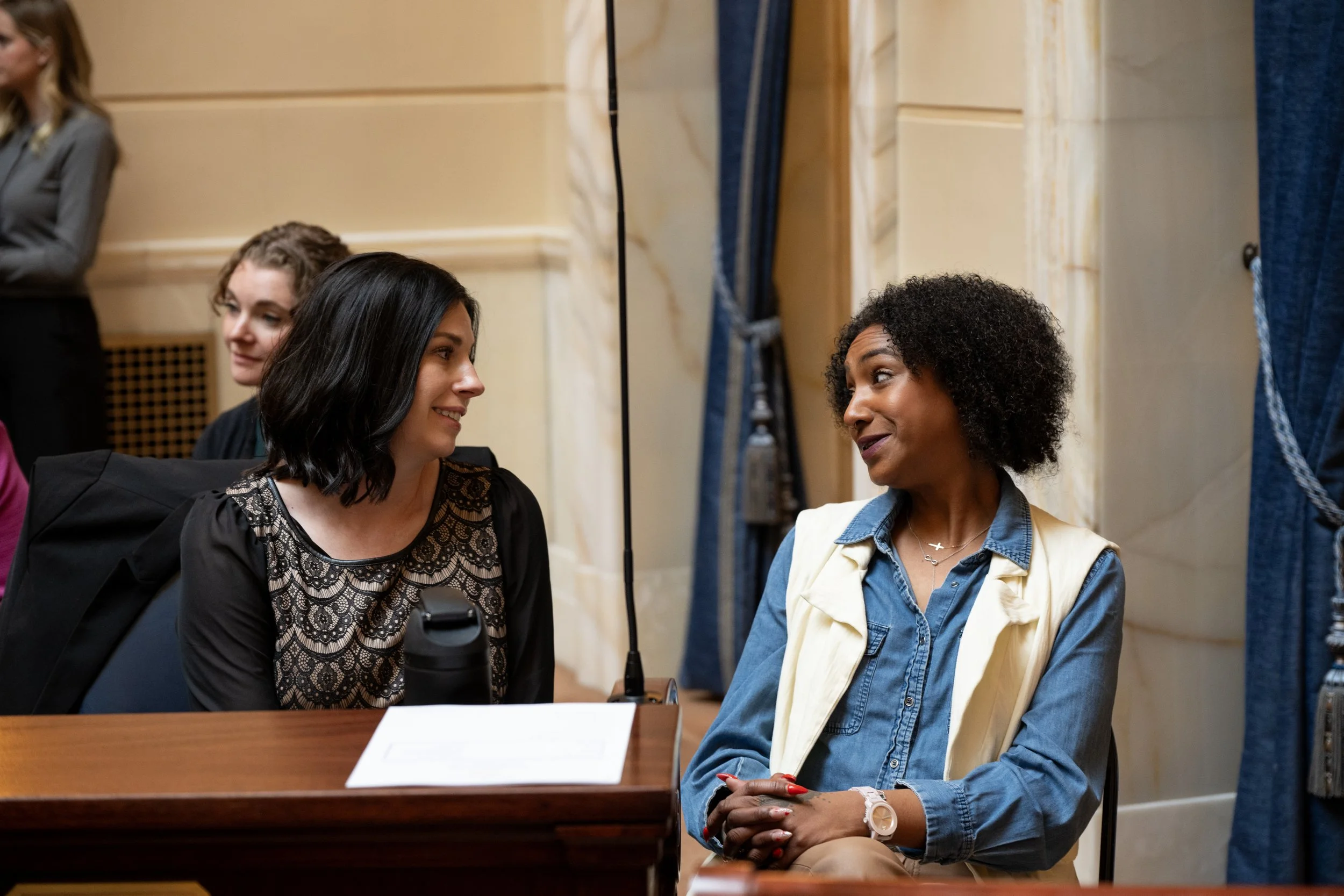 Two women are sitting and engaging in conversation, smiling at each other. One has black hair black lace top, the other has curly black hair dressed in a denim shirt and vest. They are in a formal setting, with a beige wall and blue curtains in the background.