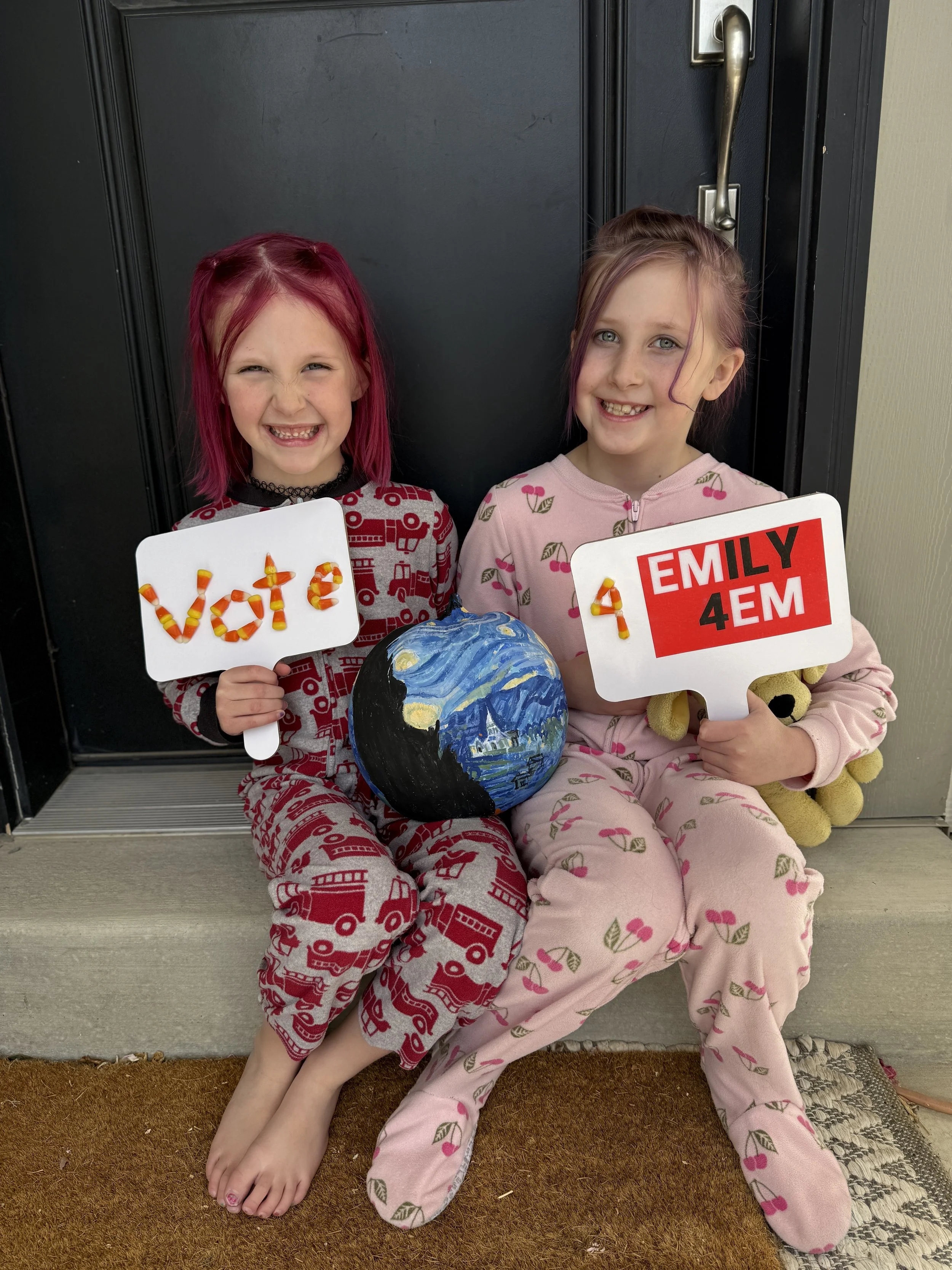Two young girls in pajamas are sitting on a doormat in front of a black door. They are holding campaign signs and a pumpkin painted to resemble Van Gogh's Starry Night painting.