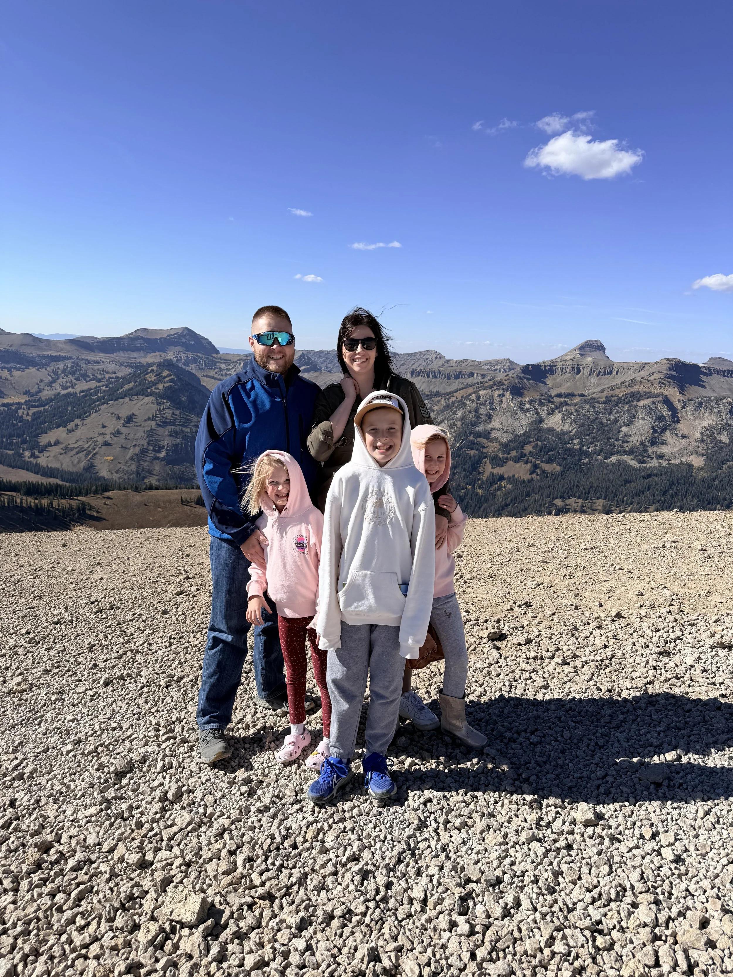 Family of five posing on rocky terrain with mountains and blue sky in the background.