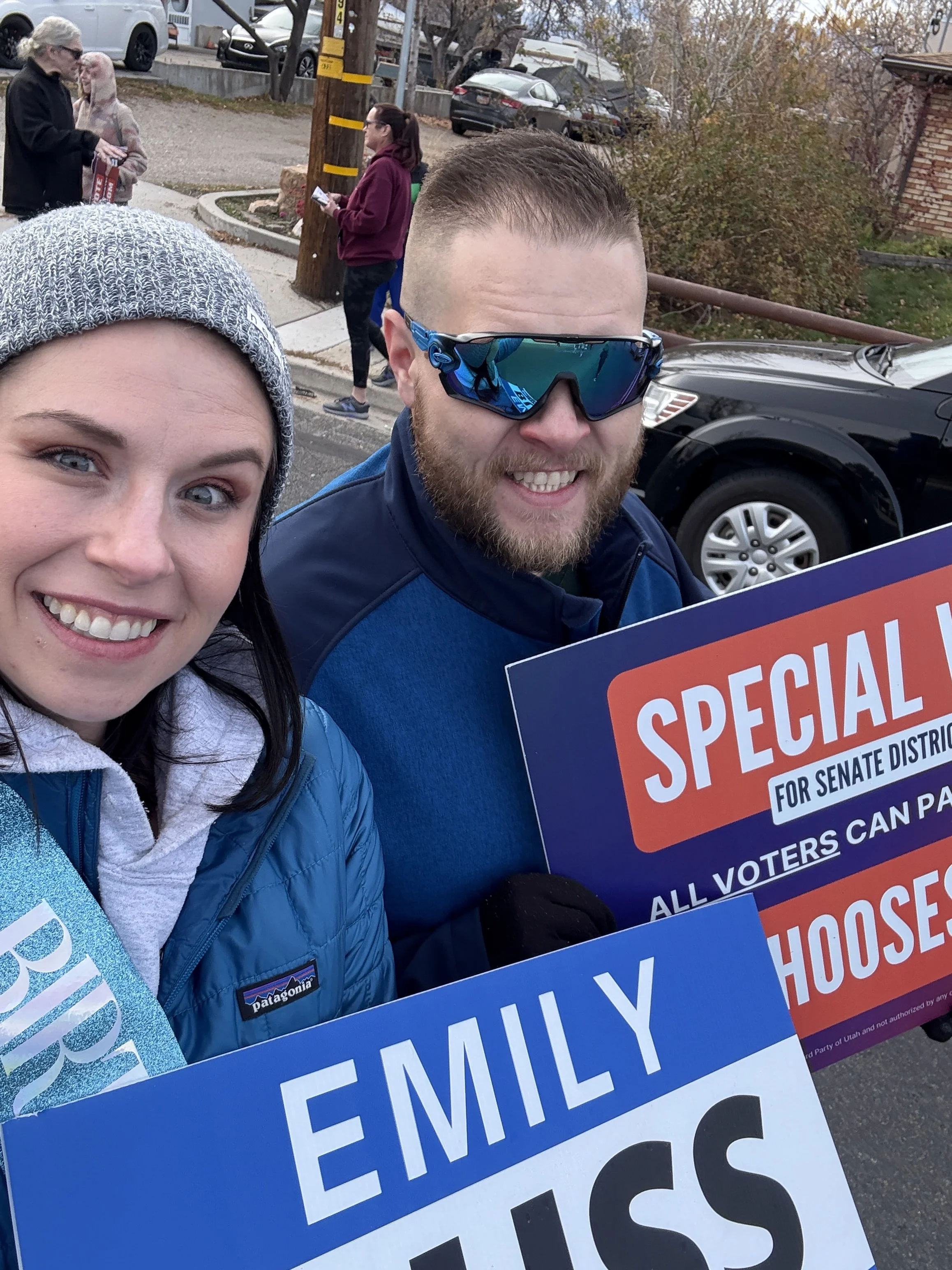 Two smiling adults taking a selfie outdoors during a political parade. Senator Emily Buss, wears a gray knit hat and blue jacket, the man wears sunglasses and a blue jacket. They are holding signs supporting Candidate Emily Buss, and voting.