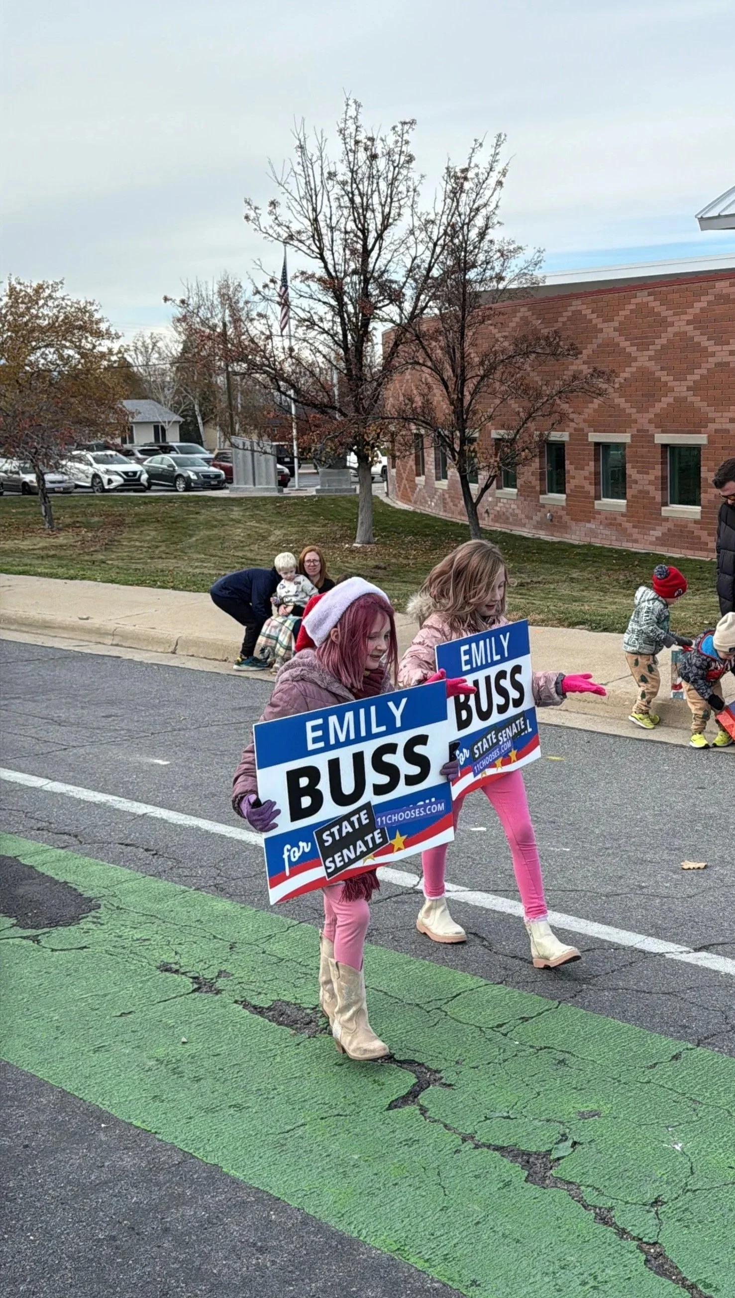 Children holding campaign signs for Emily Buss during a parade on a street with a brick building in the background.
